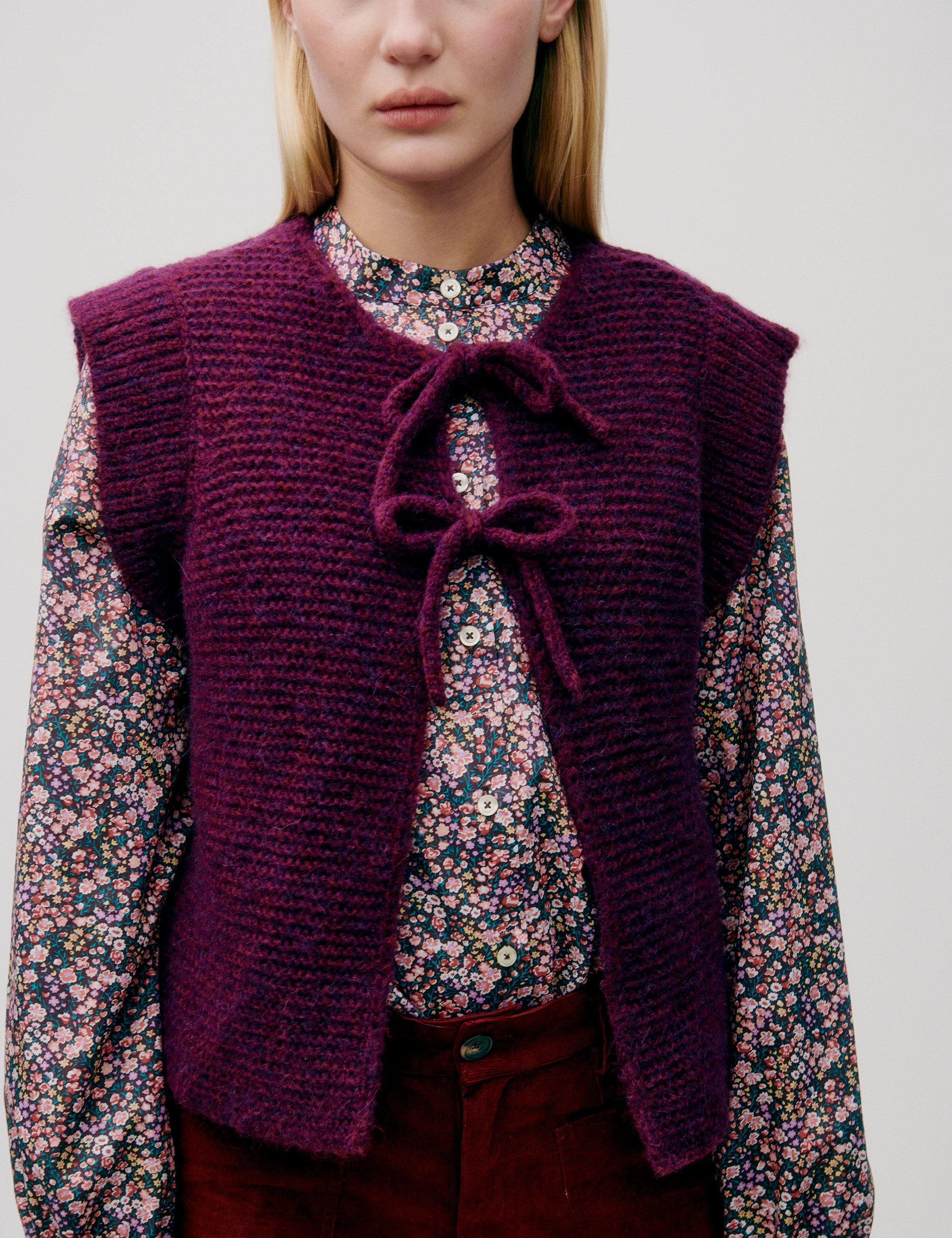 Woman in pink and blue flower patterned shirt with long sleeves, dark pink knitted wool vest and red pants