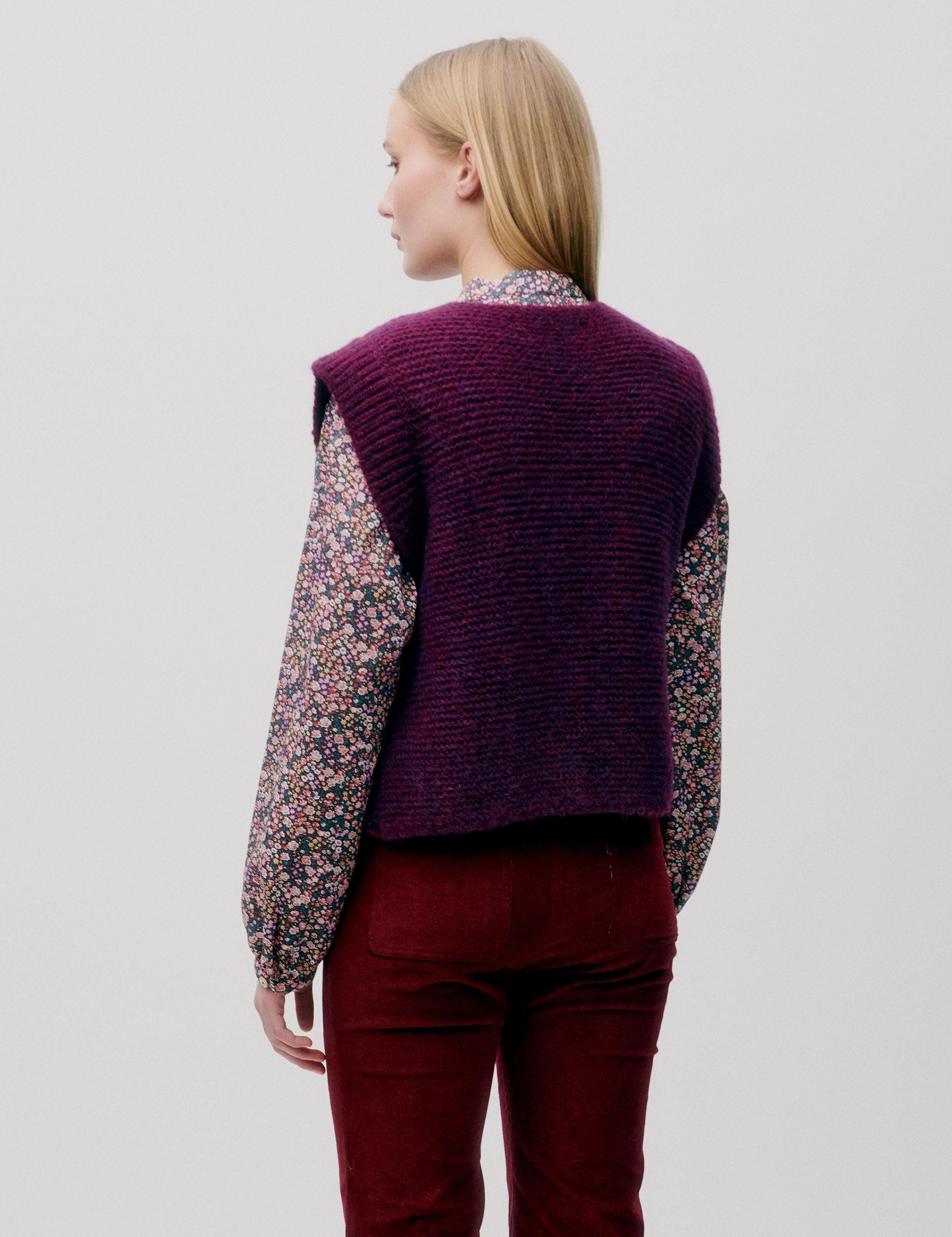 Woman in pink and blue flower patterned shirt with long sleeves, dark pink knitted wool vest and red pants