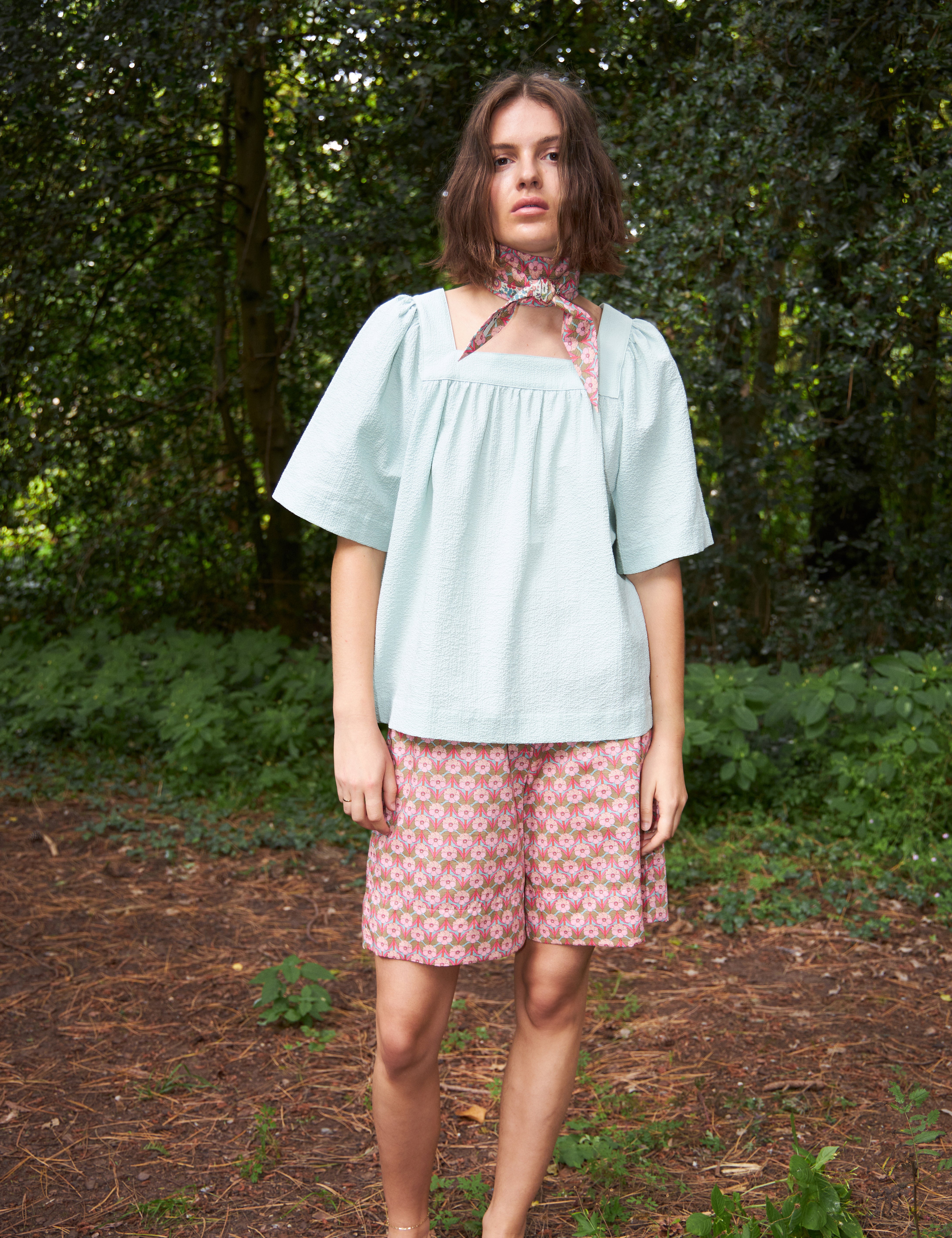 Woman in pink floral patterned shorts and light blue loose fit blouse with short sleeves and bandana scarf around neck