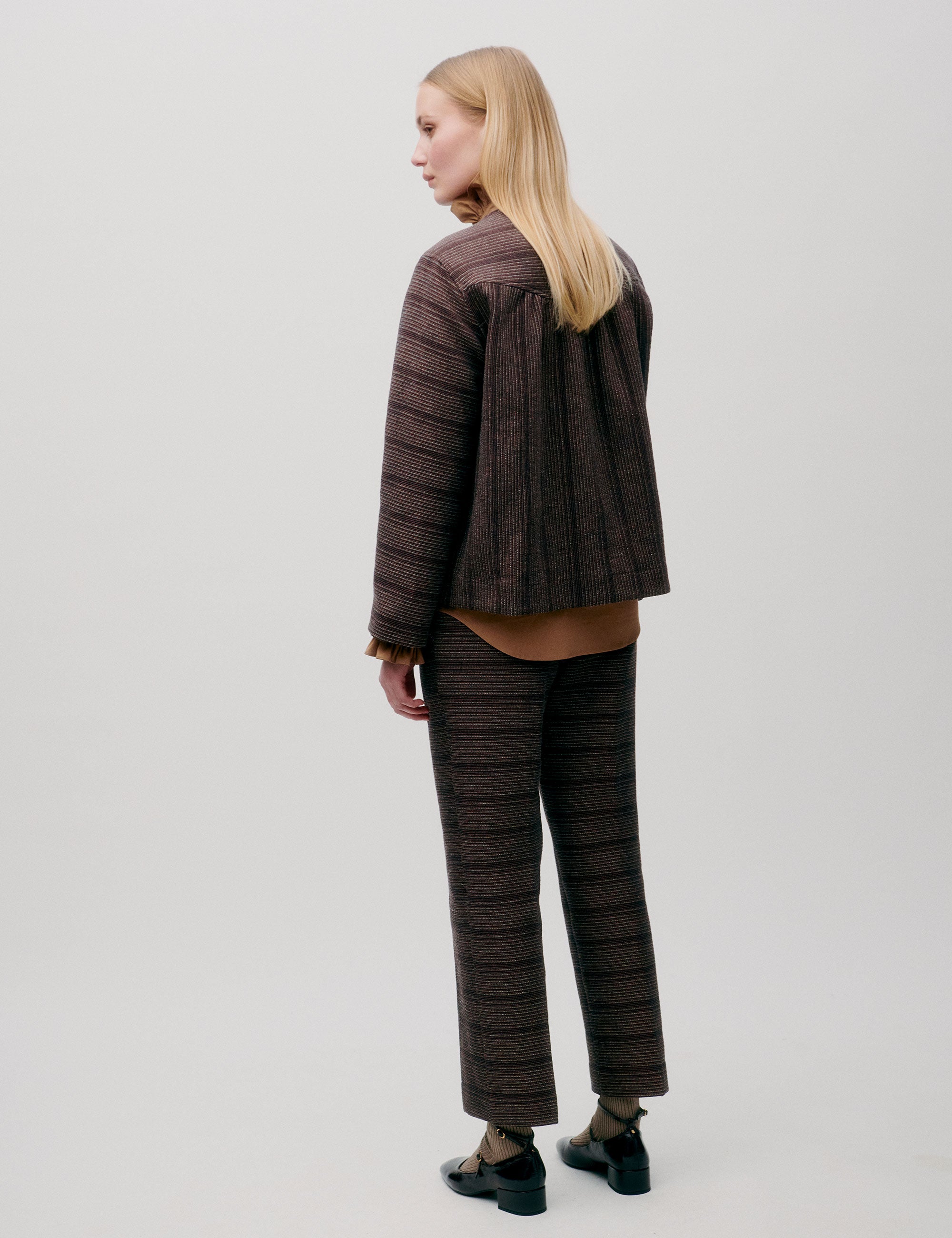Woman in brown striped jacket with pockets and large buttons, matching straight pants and brown shirt with frilled collar
