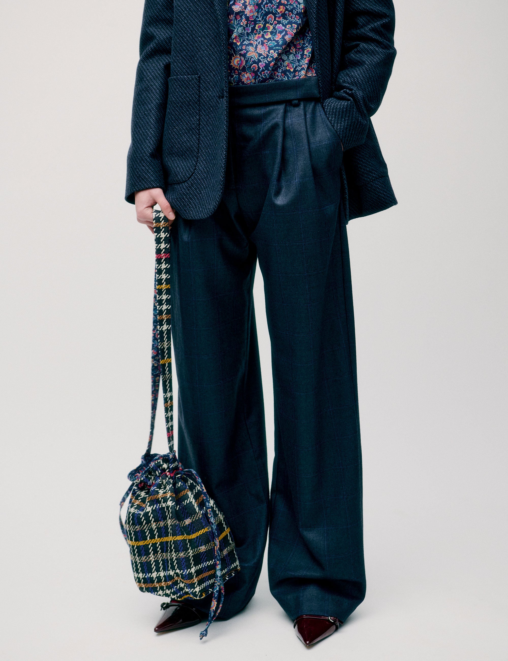 Woman wearing dark wide-leg trousers styled with a printed blouse and blazer, holding a checkered bag