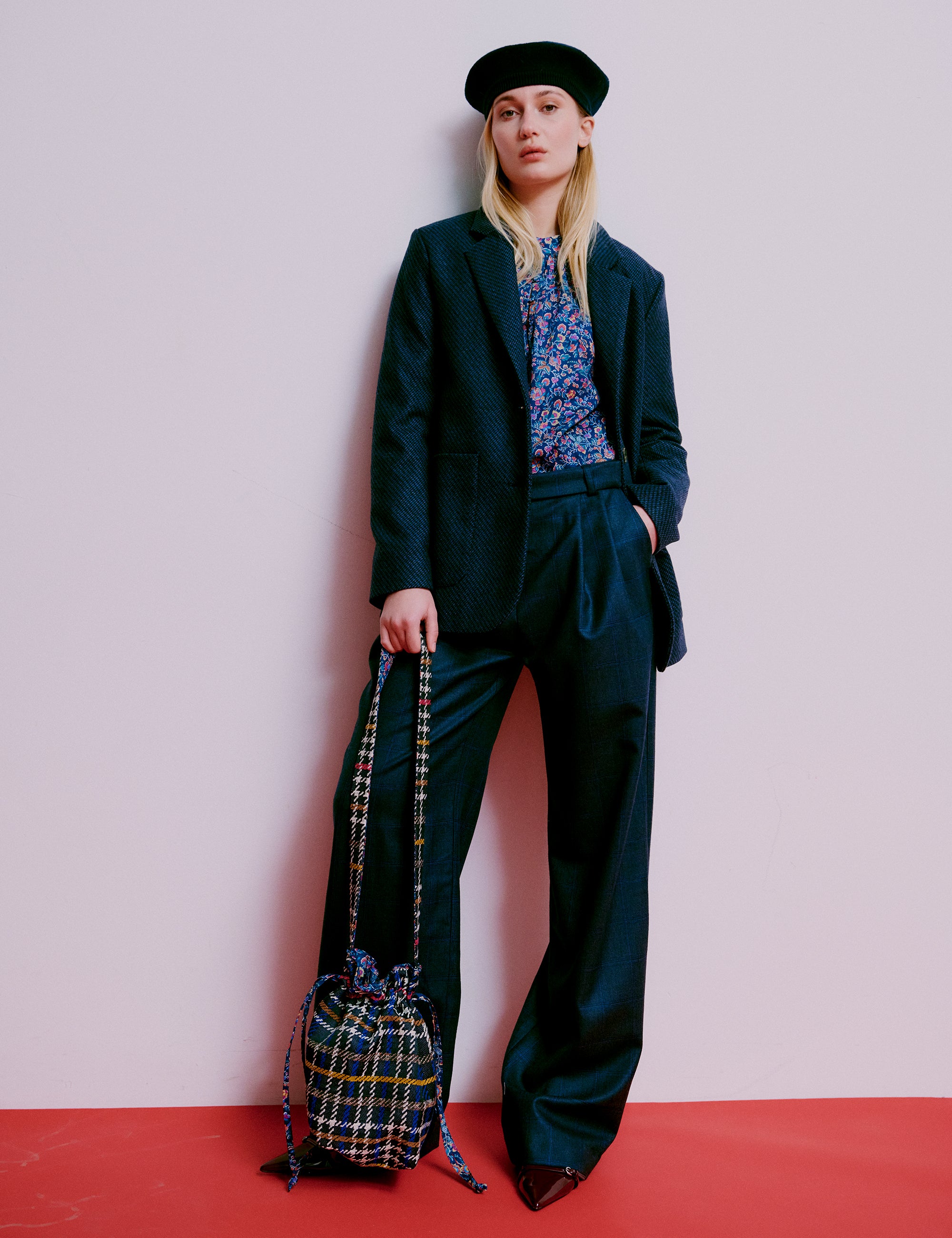 Woman wearing a dark suit with wide-leg pants and a beret, holding a plaid bag on a red floor against a white wall.