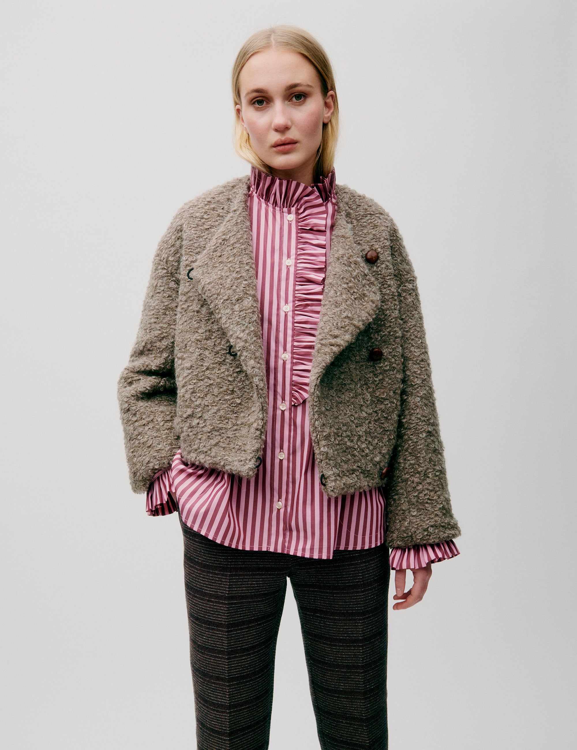 Woman in short wool jacket with assymetric design and a bubbly texture, a pink striped shirt with frilled chest and brown striped pants