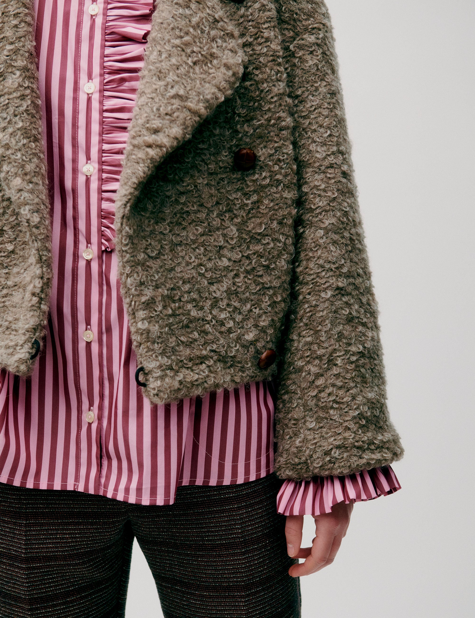 Woman in short wool jacket with assymetric design and a bubbly texture and a pink striped shirt with frilled chest