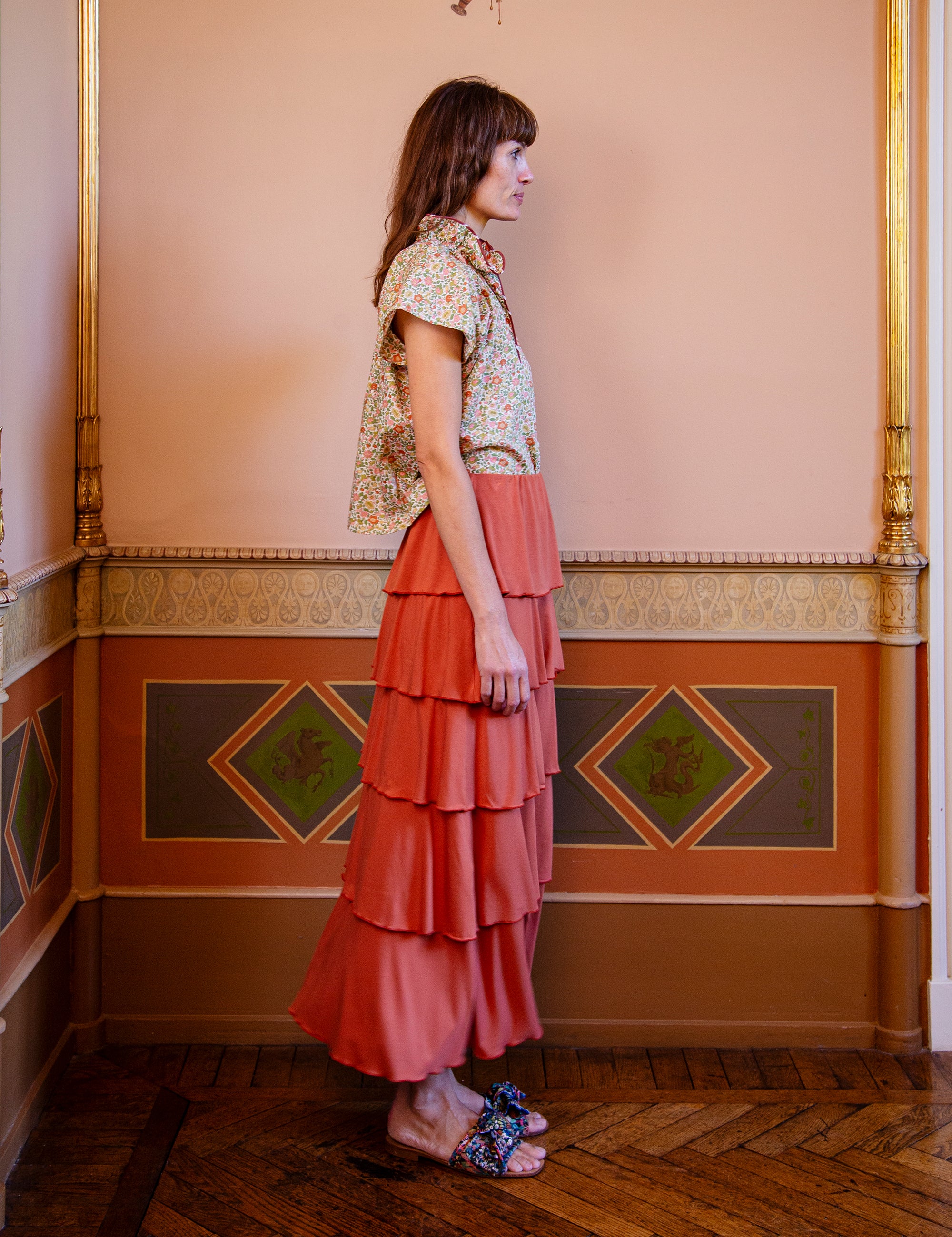 Woman in a floral blouse and coral tiered skirt standing in an ornate room with decorative walls.
