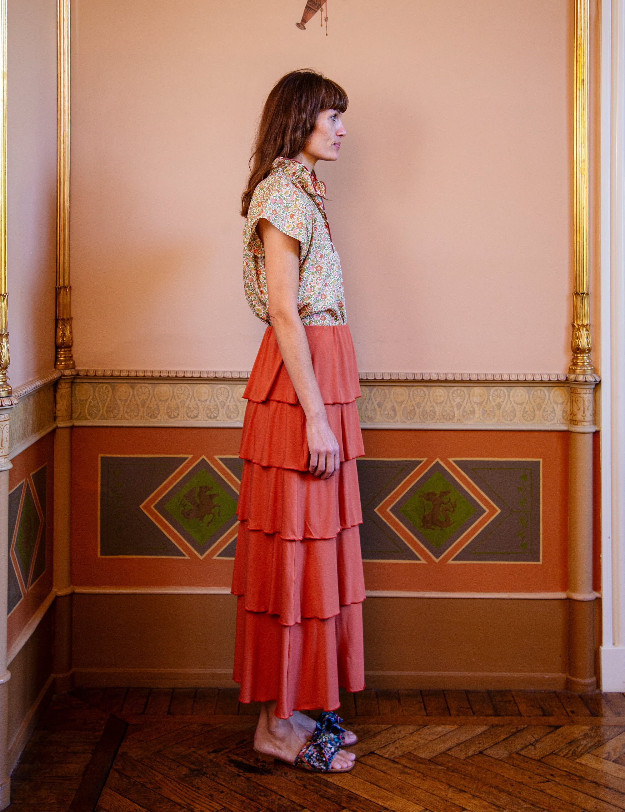 Woman in a floral top and tiered coral skirt standing in an ornately decorated room.