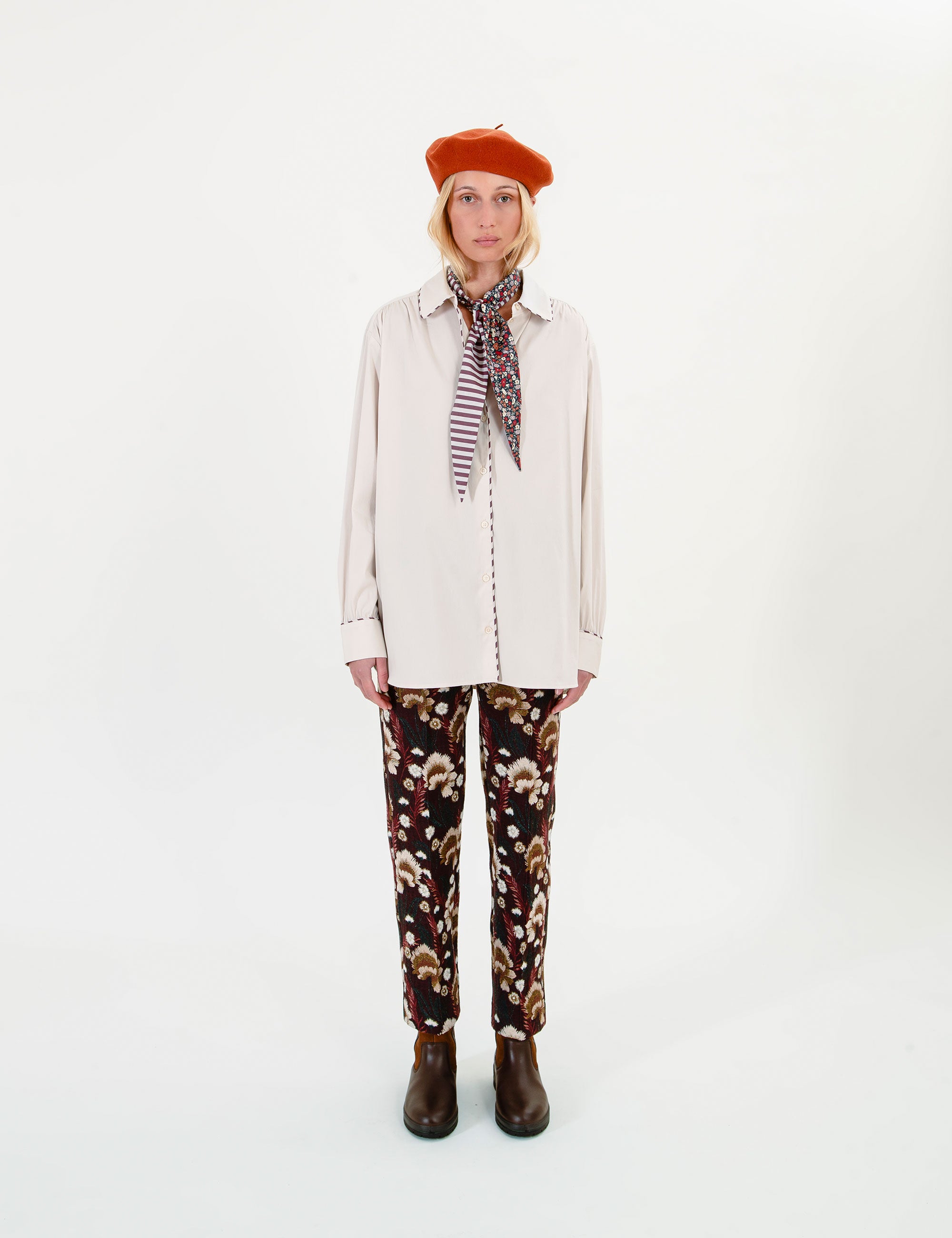 Woman wearing a beige shirt, orange beret, striped and floral scarf and brown floral pants on a white background