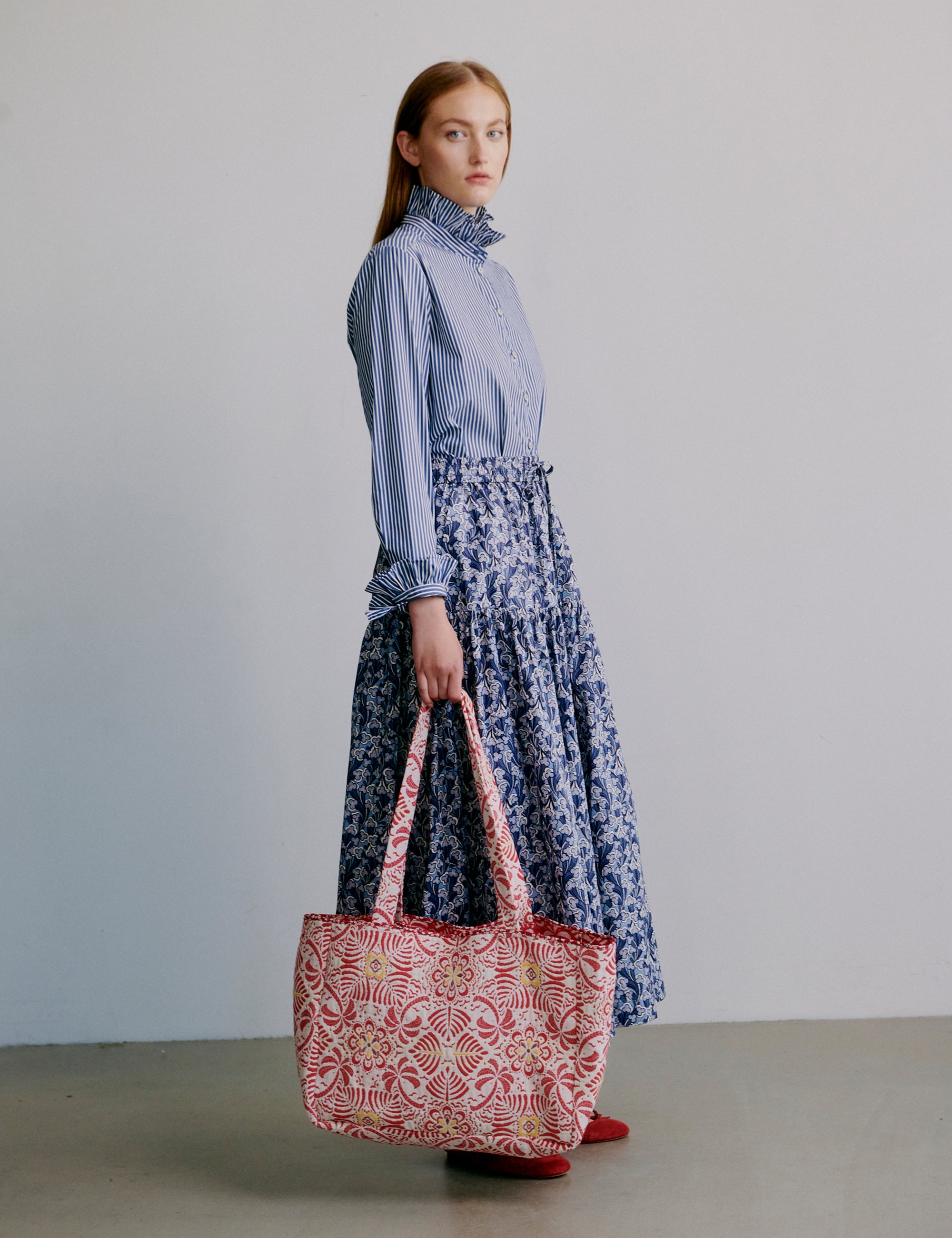 Woman wearing a blue striped shirt and blue floral skirt holding a red patterned bag against a plain background