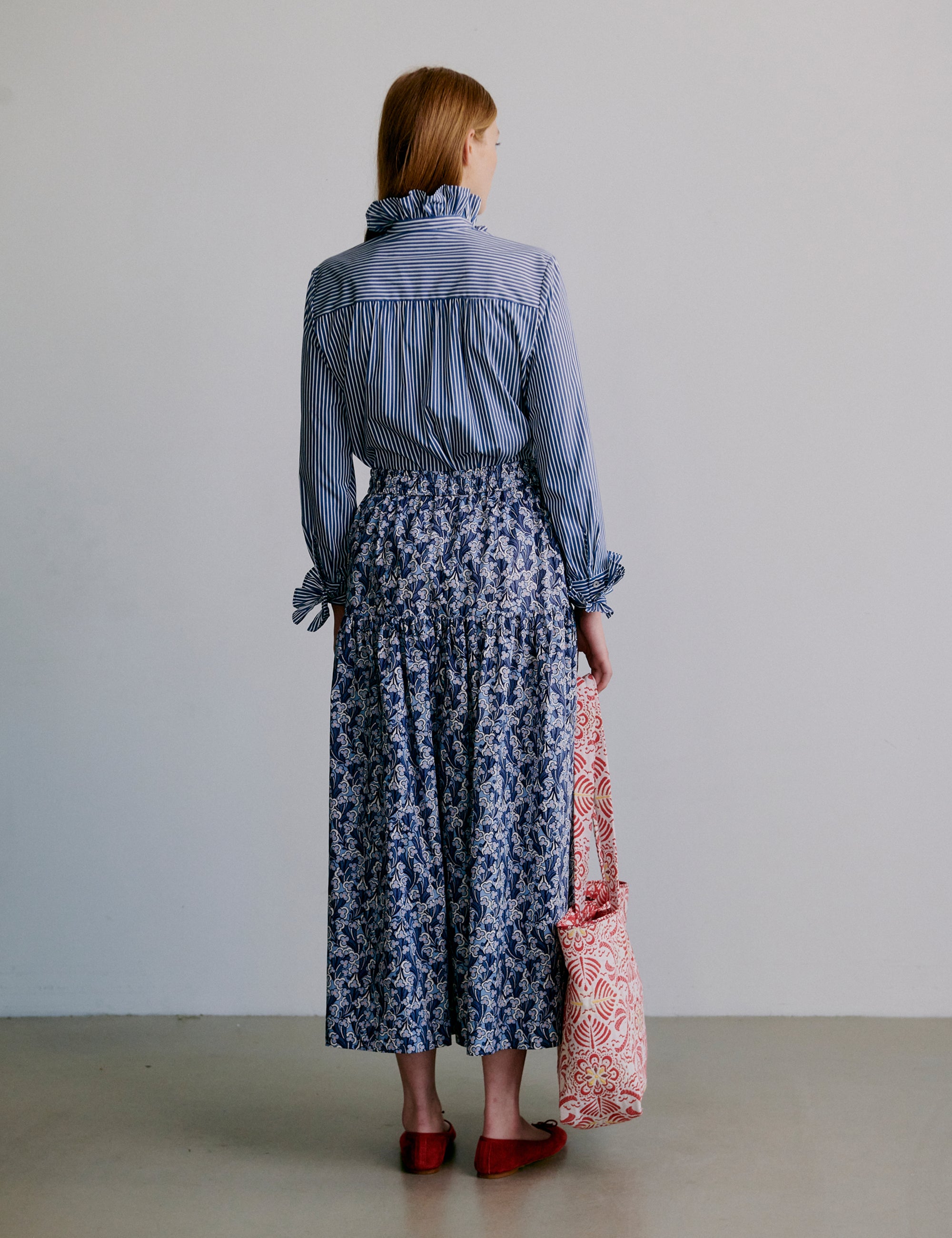Woman wearing a blue striped shirt and blue floral skirt holding a red patterned bag against a plain background