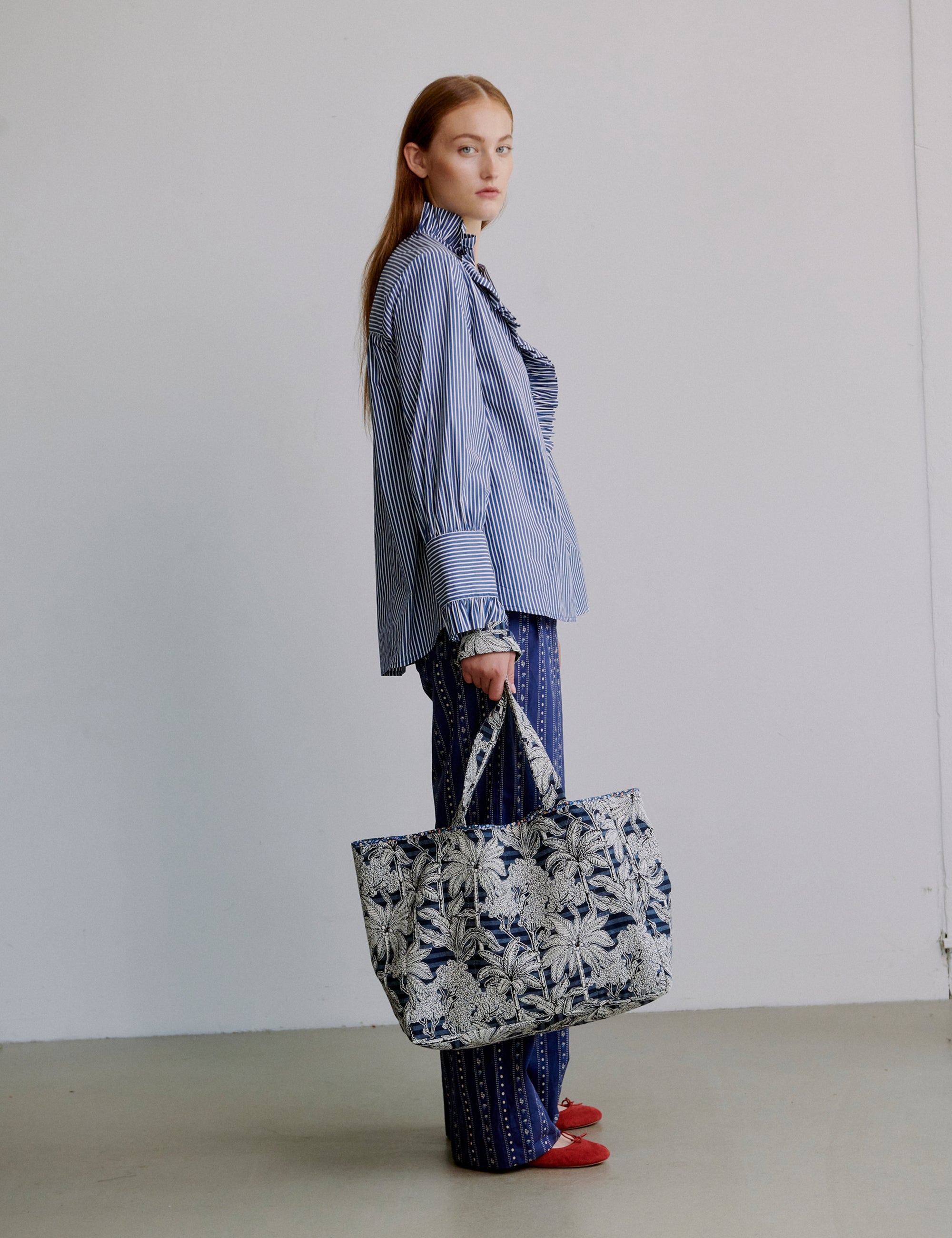 Woman wearing a blue and white striped shirt with frilled collar and cuffs, blue patterned pants and floral patterned tote bag
