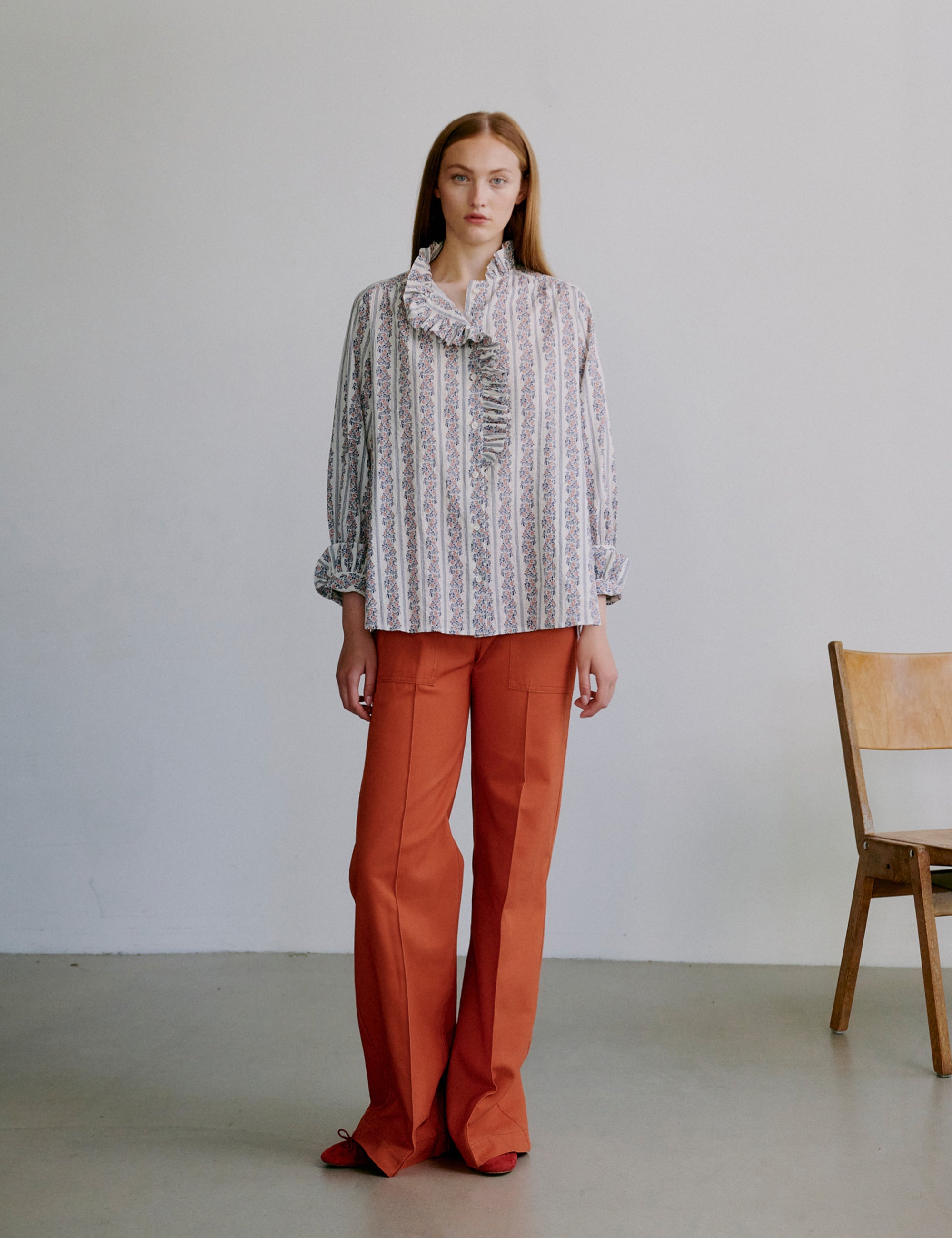 Woman wearing a floral patterned blouse and orange pants standing against a plain background with a wooden chair.