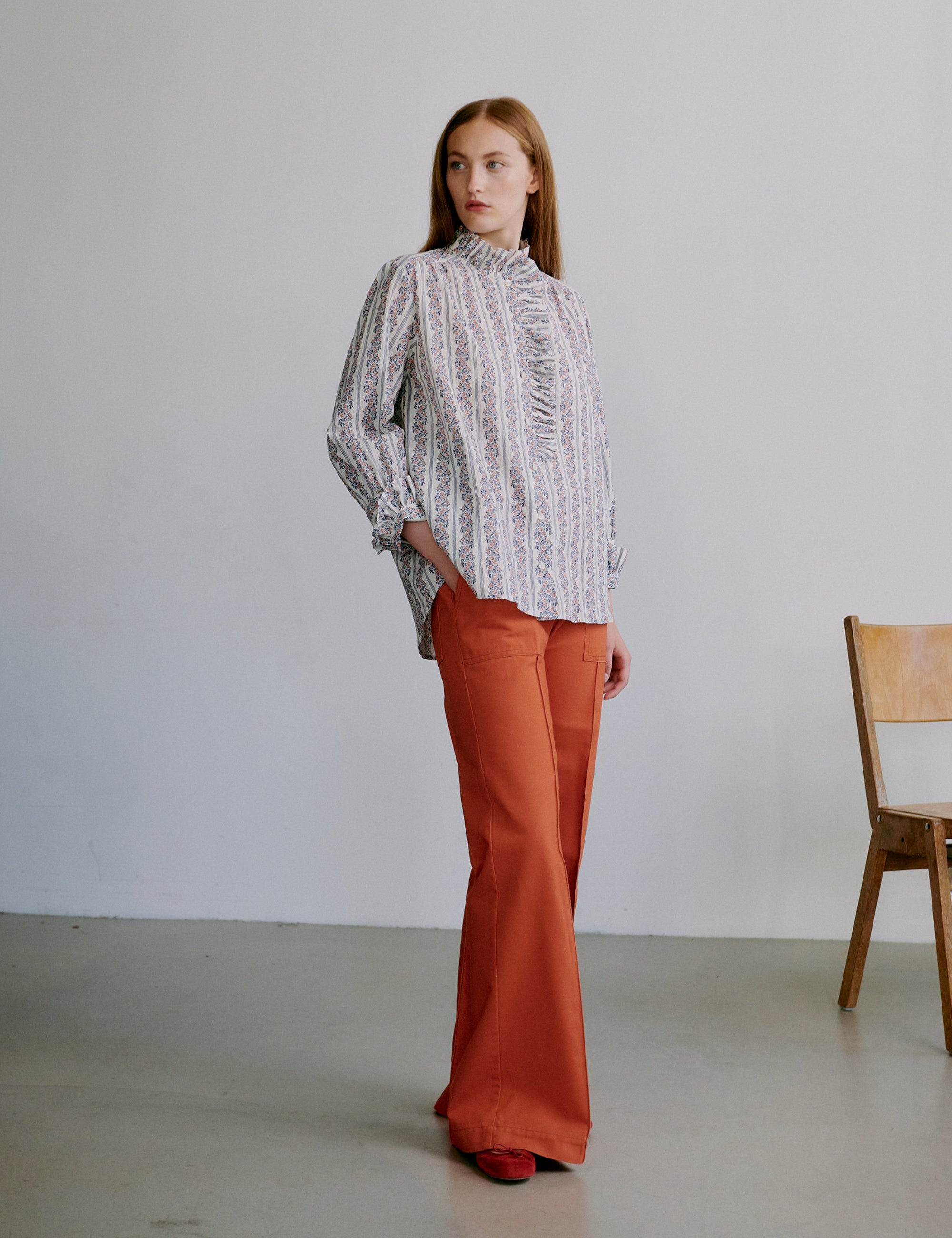 Kvinna i blommig blus med volanger och orange byxor mot neutral bakgrund med trästol