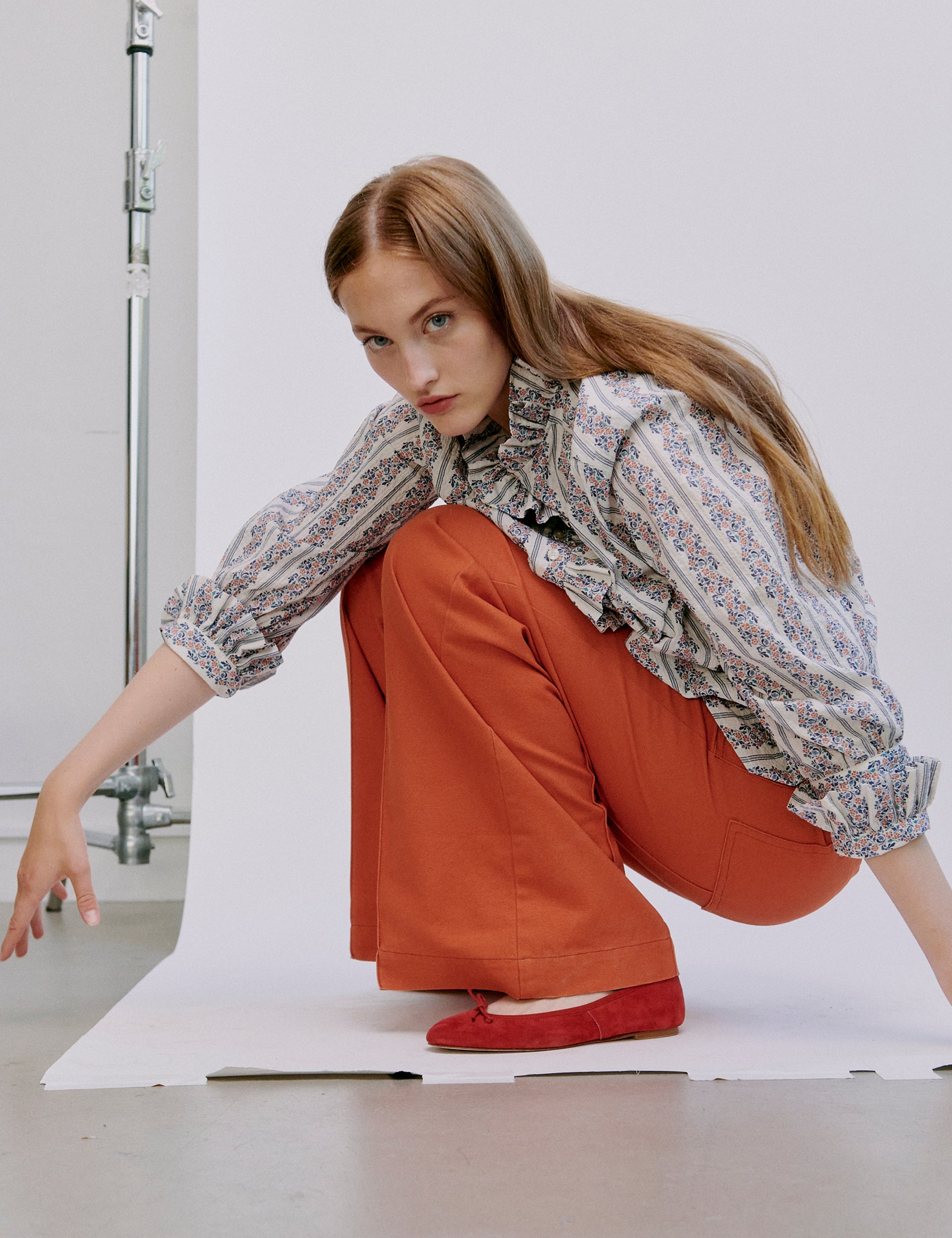 Woman sitting on a white floor wearing a patterned blouse and orange pants.