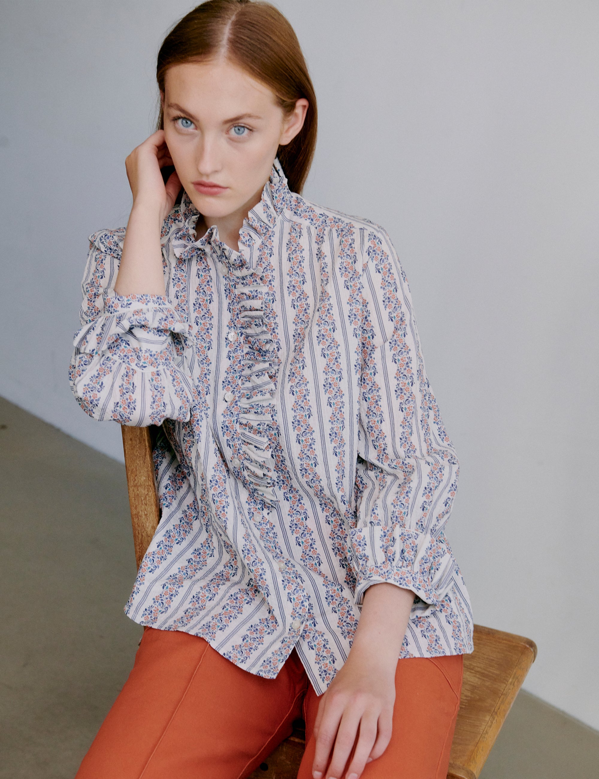 Woman wearing a floral patterned blouse and orange pants sitting on a wooden chair.