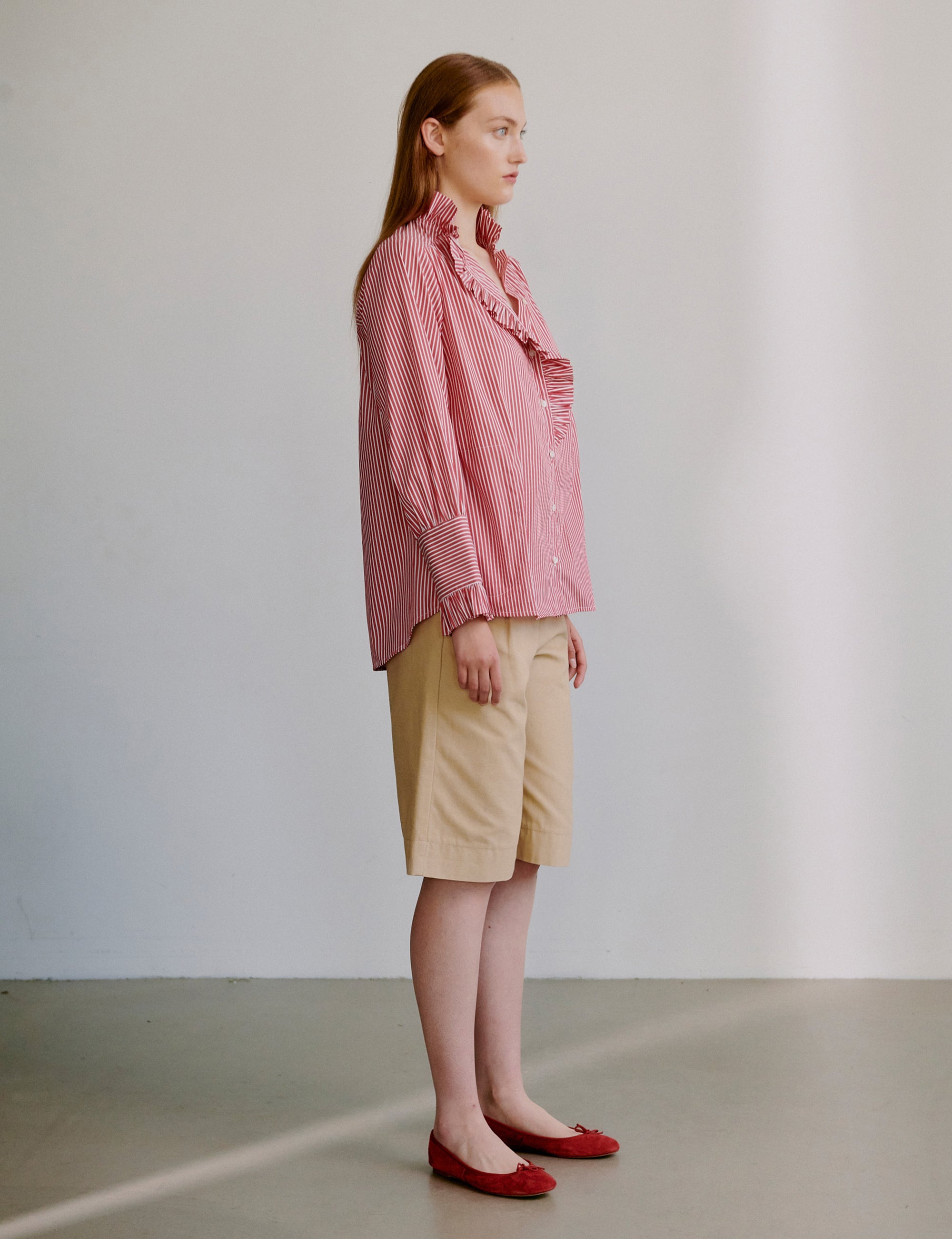 Woman wearing a pink striped shirt with frilled details and beige shorts against a plain background
