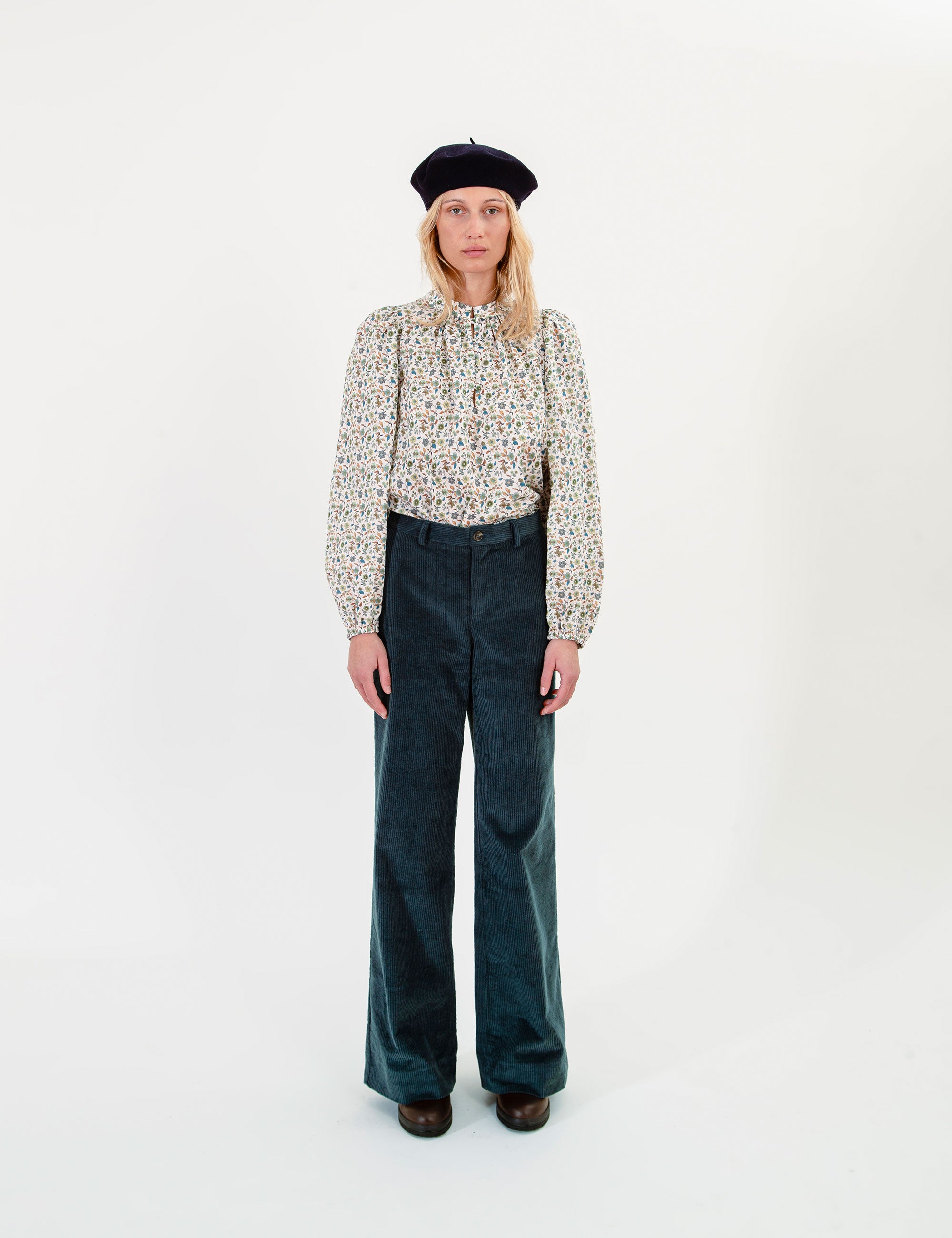 Woman wearing a patterned blouse and dark corduroy pants on a white background
