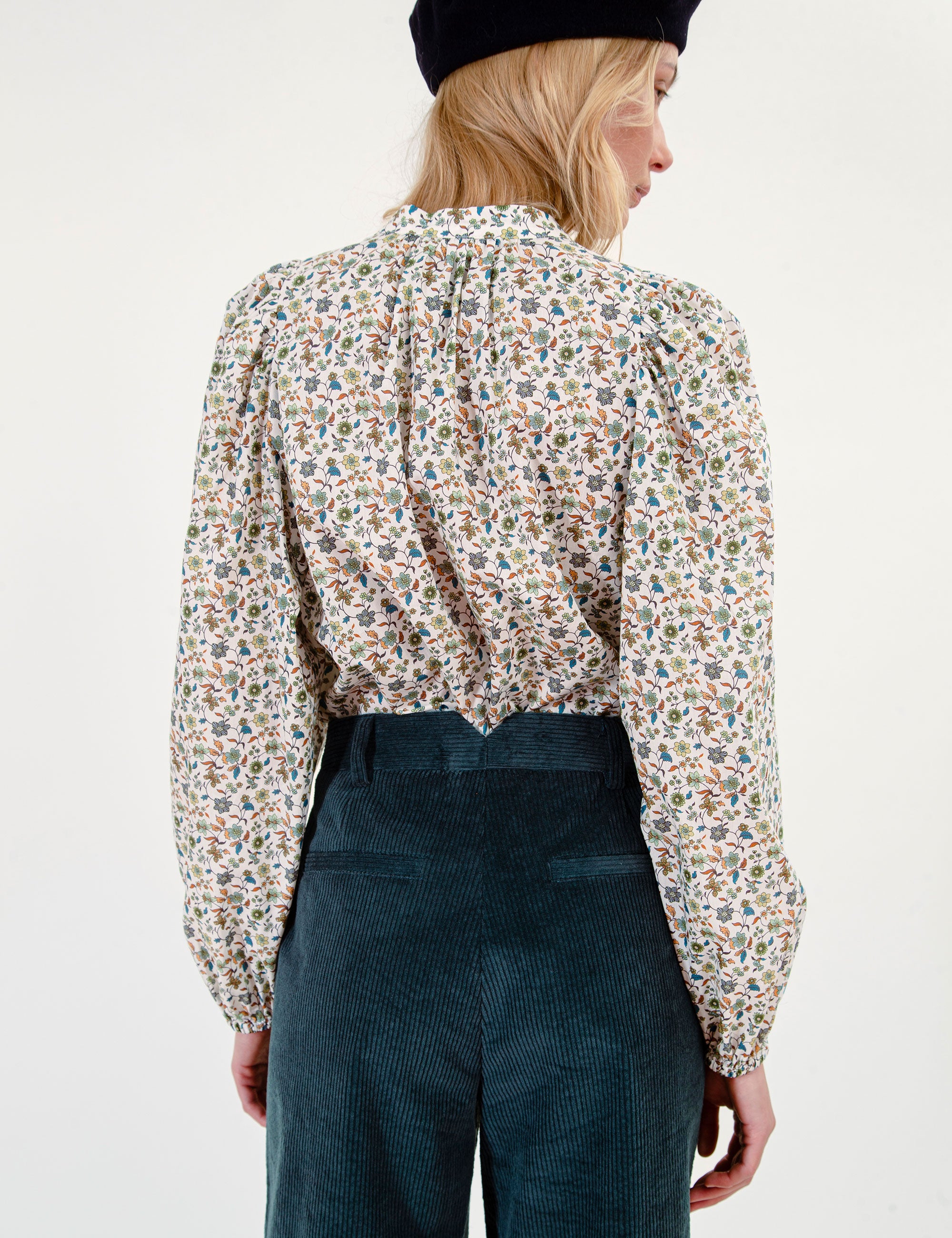 Woman wearing a floral patterned blouse and dark corduroy pants on a white background