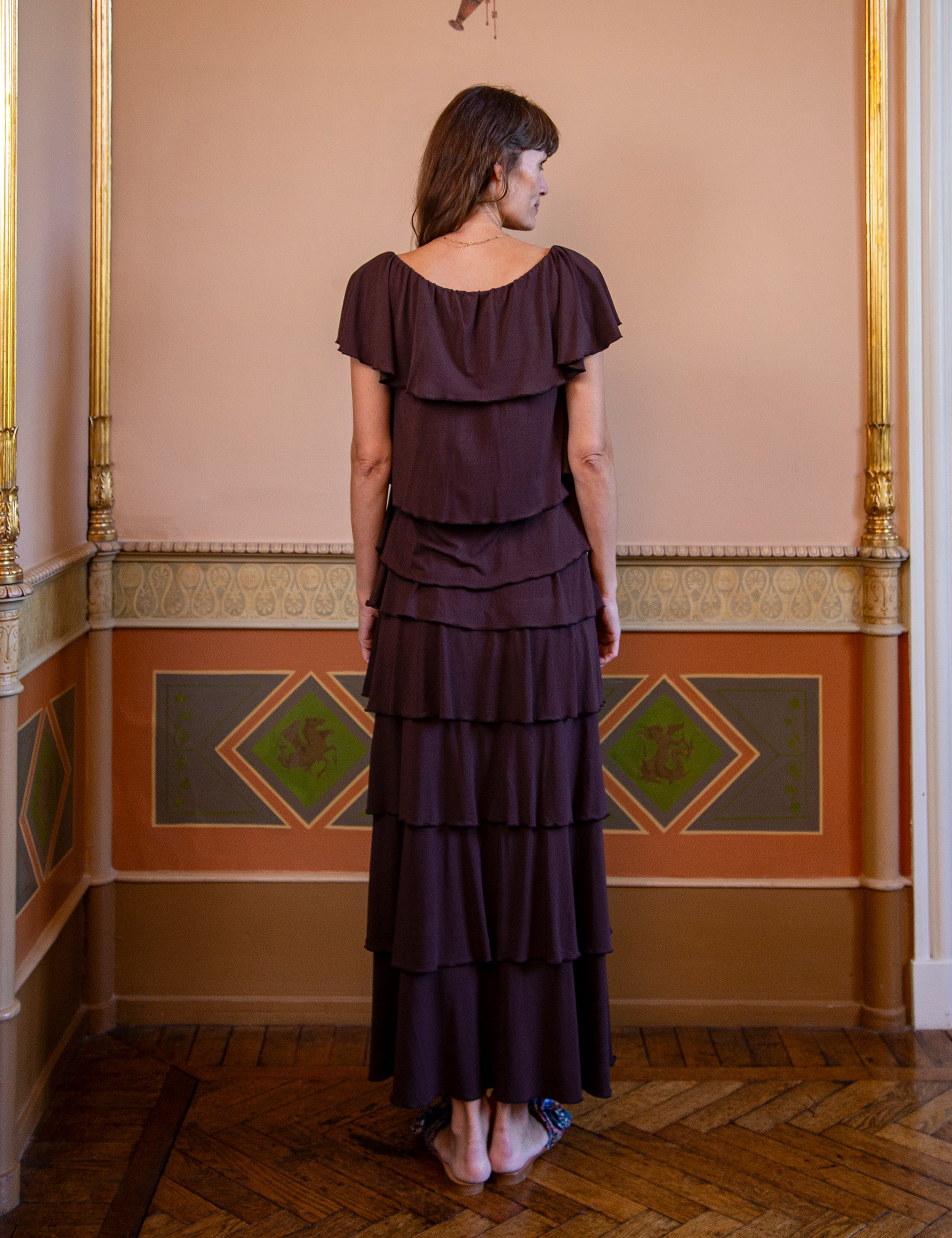 Woman wearing a dark brown tiered skirt and matching top standing in an ornate room with decorative walls.
