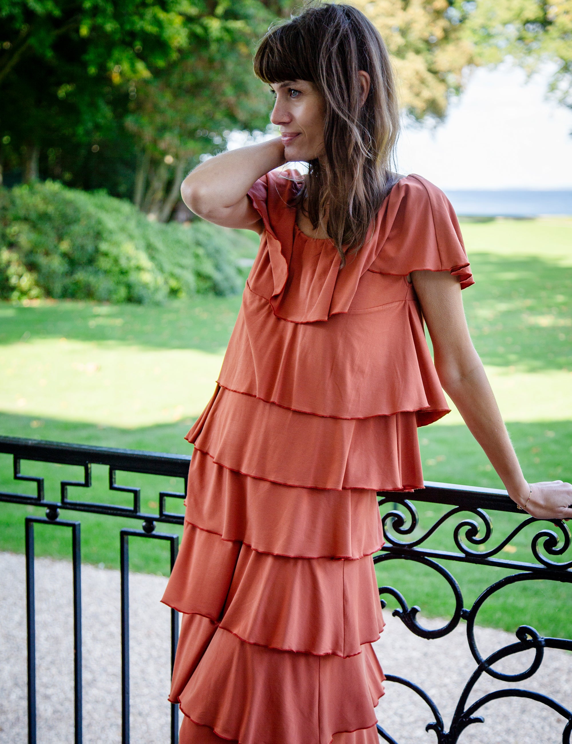 Woman wearing a layered orange top and skirt standing outdoors with greenery in the background