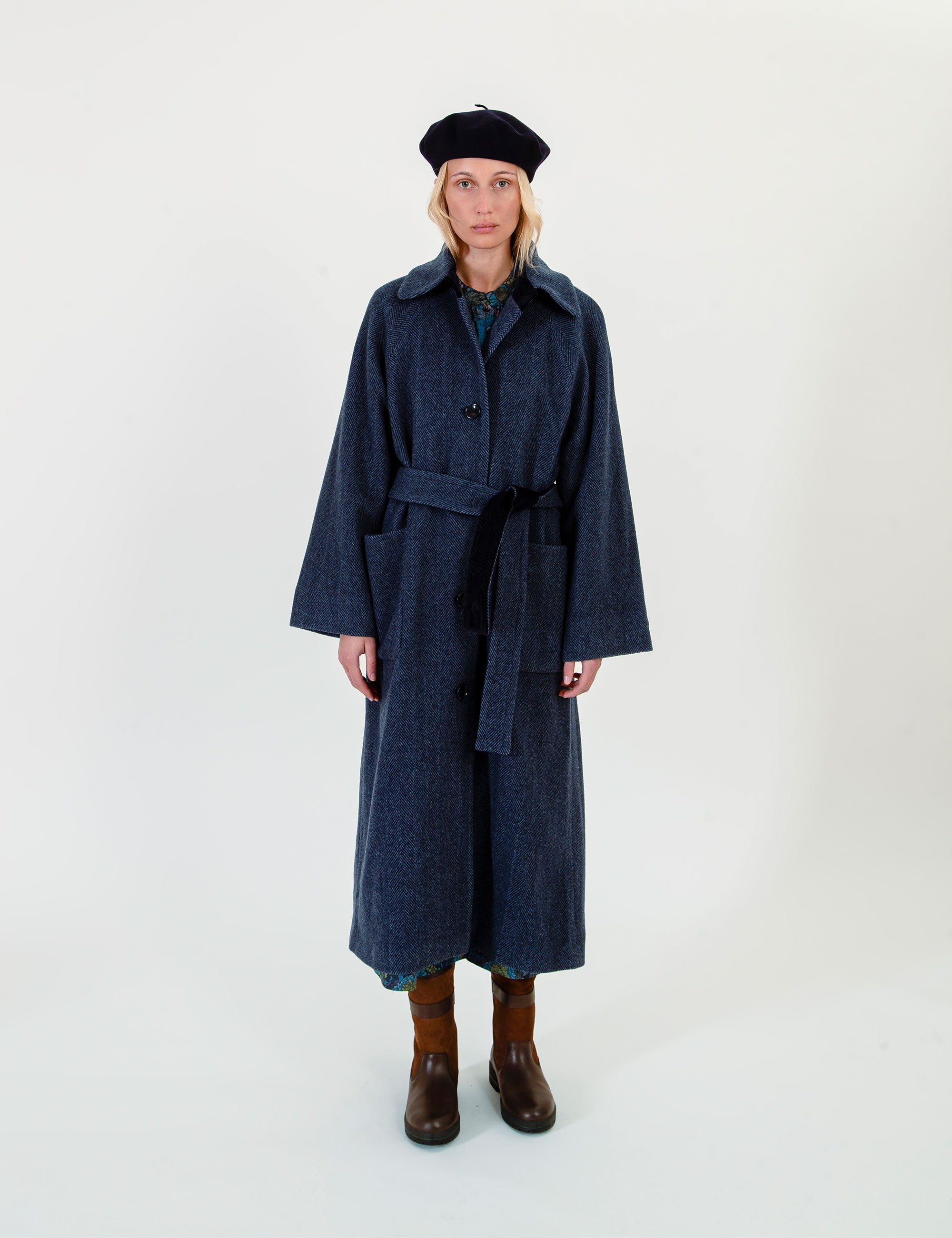 Woman in a long navy blue wool coat with button details, belt and pockets on a light gray background