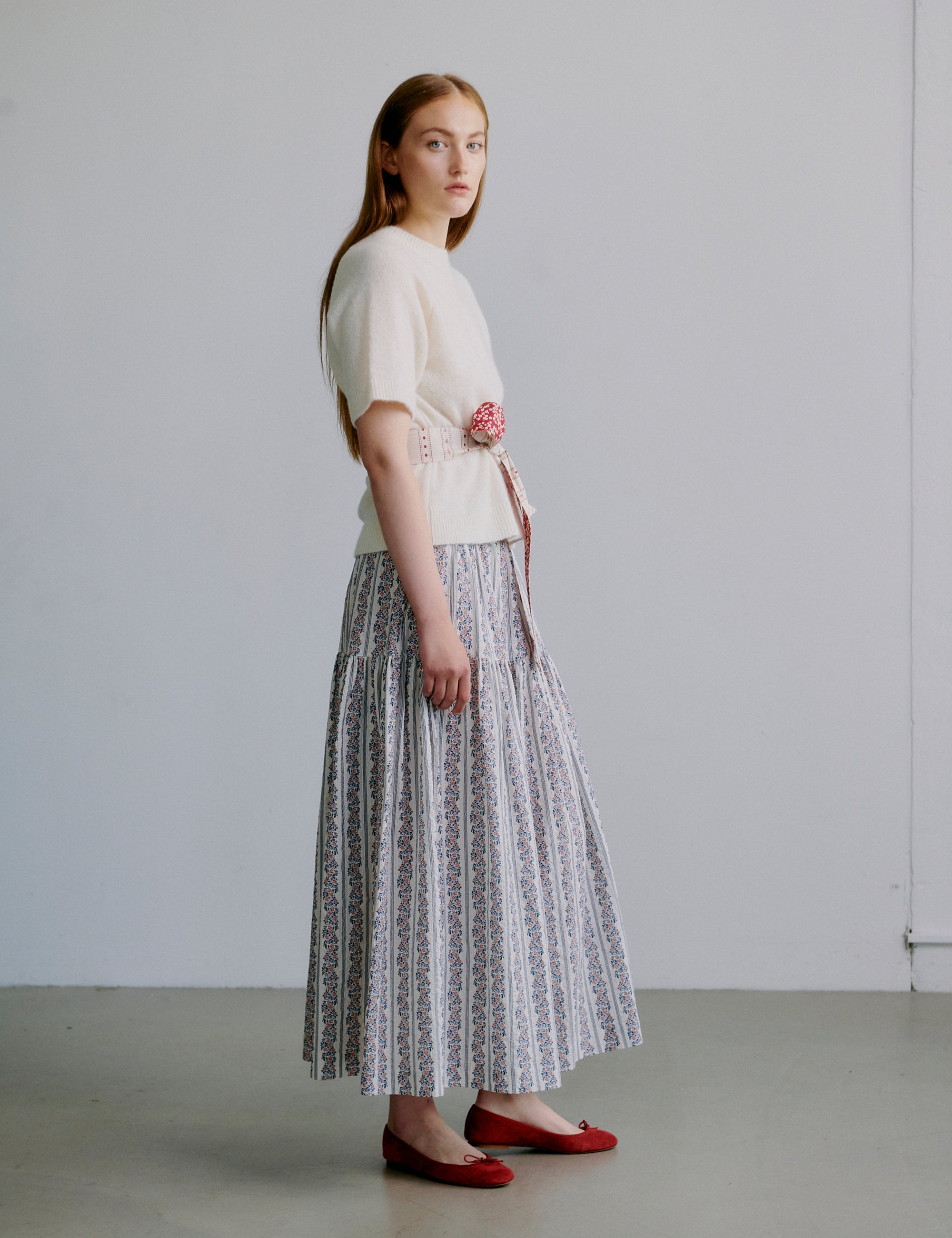 Woman wearing a white short sleeved knit top and floral patterned skirt with red shoes against a plain background