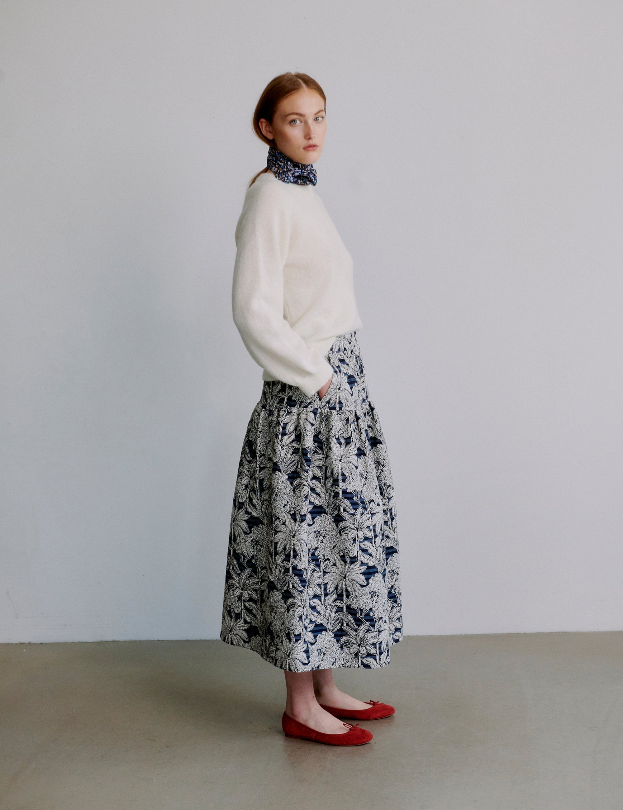 Woman wearing a white sweater and floral skirt with red shoes against a plain background