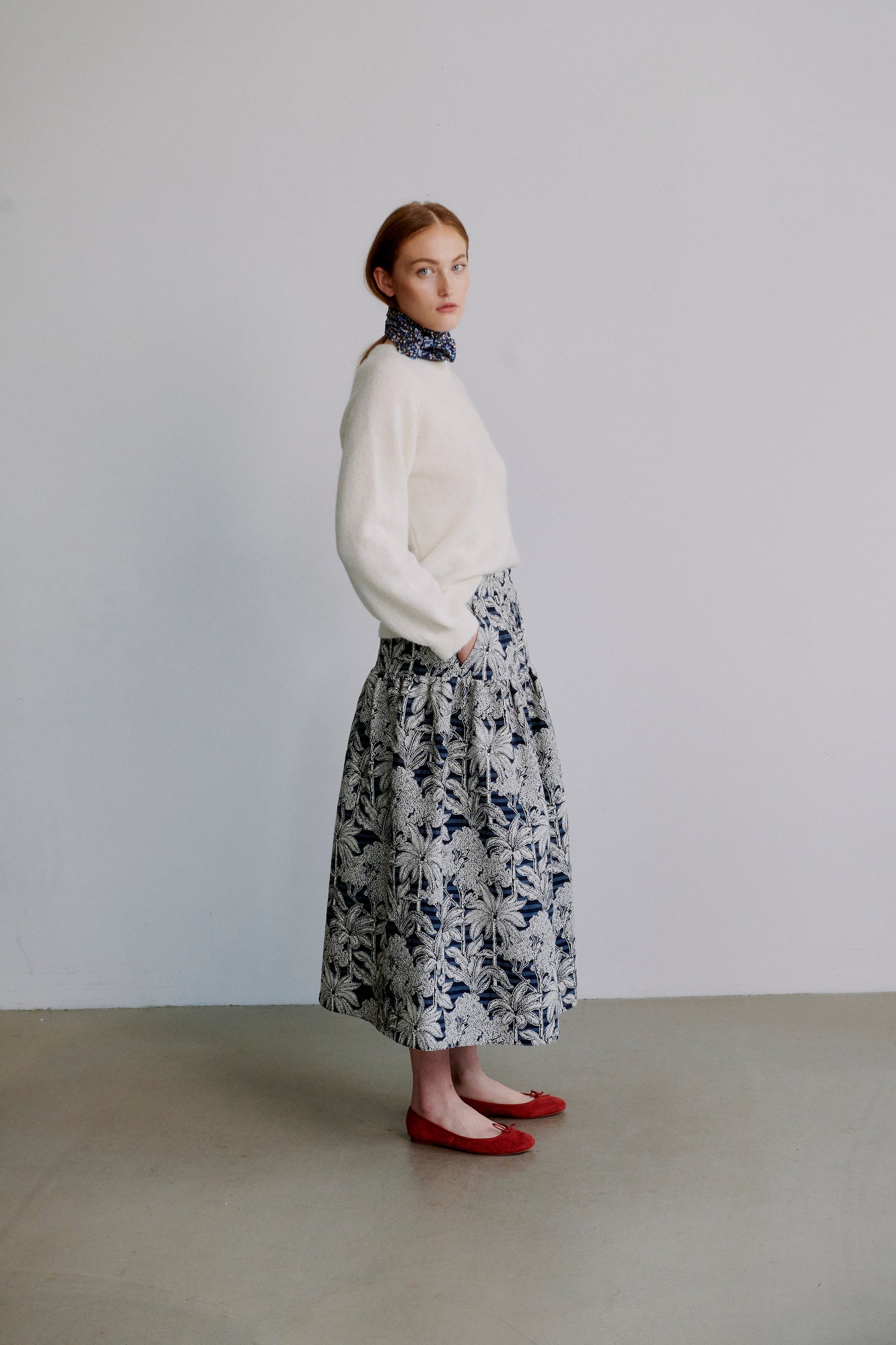 Woman wearing a cream colored long sleevew knit sweater and blue floral skirt with a blue patterned scarf against a plain background