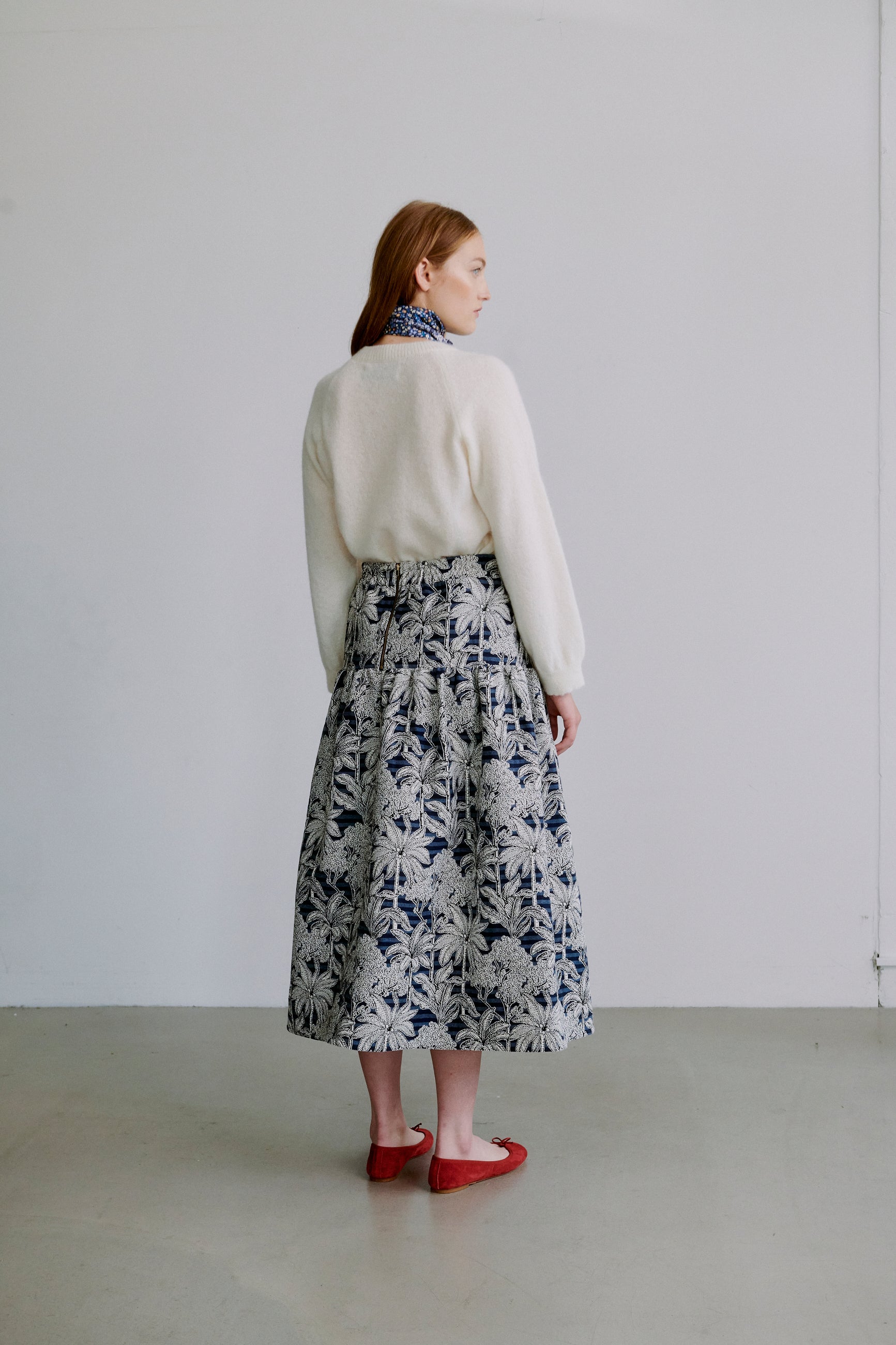 Kvinde iført en cremefarvet sweater og blomstret nederdel mod hvid baggrund