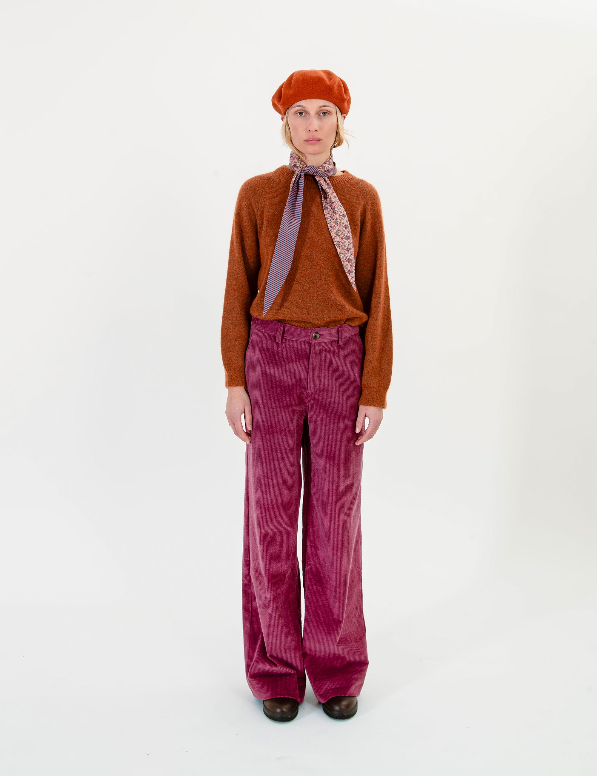 Woman wearing a brown sweater, red beret, and pink corduroy pants on a white background