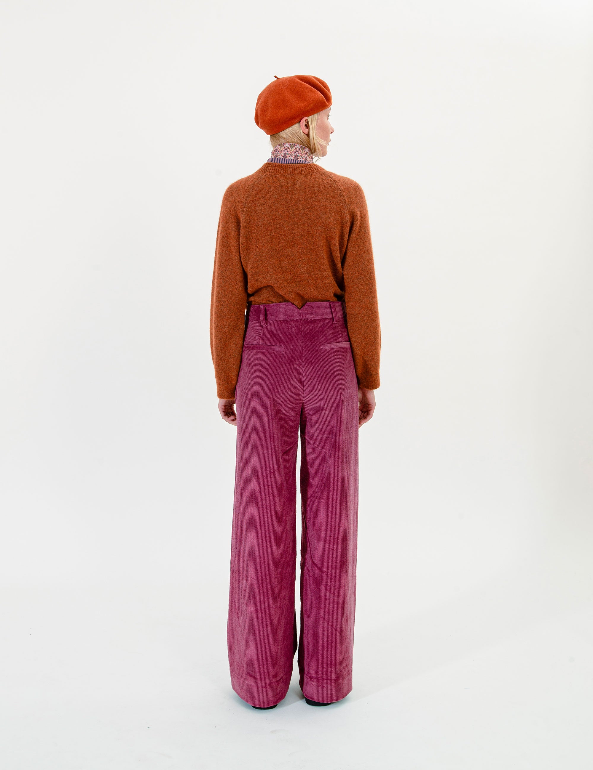 Woman wearing a brown sweater, red beret, and pink corduroy pants on a white background