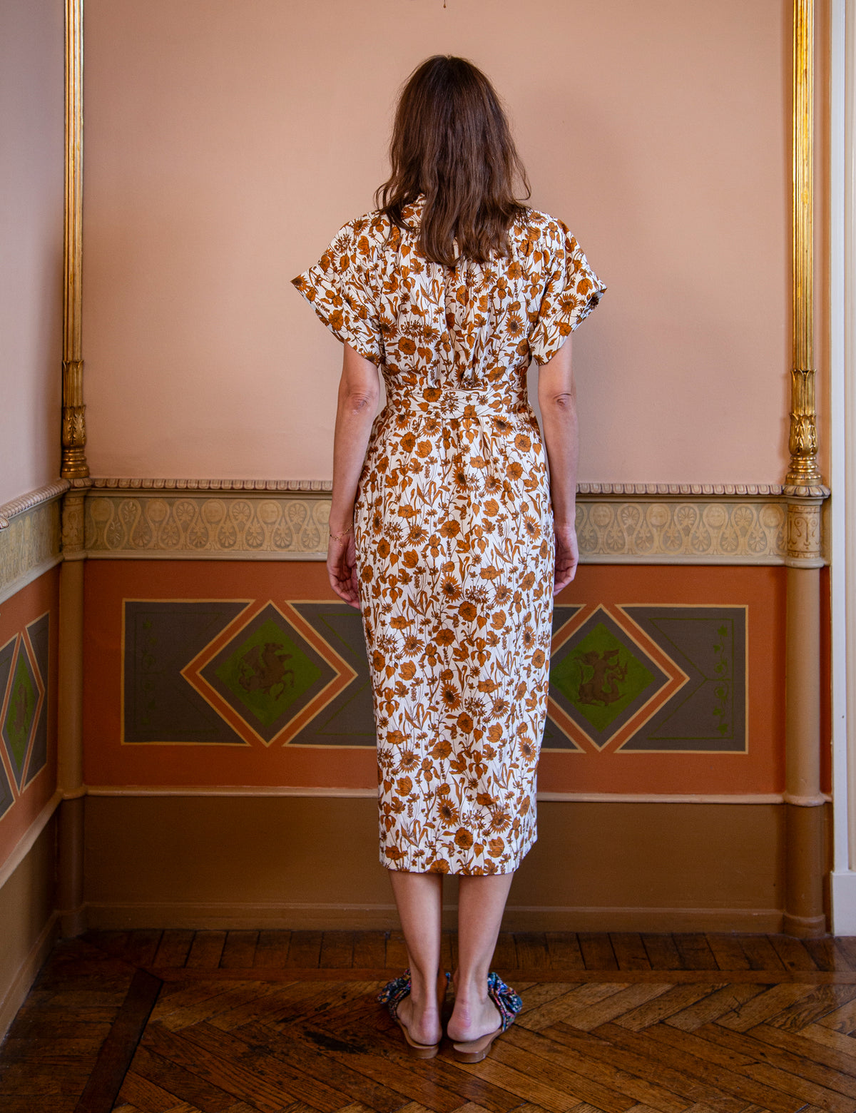 A woman viewed from behind wearing a short-sleeved midi dress with a brown and orange floral pattern on a white background, standing in an indoor room with a decorative wall and wooden floor.