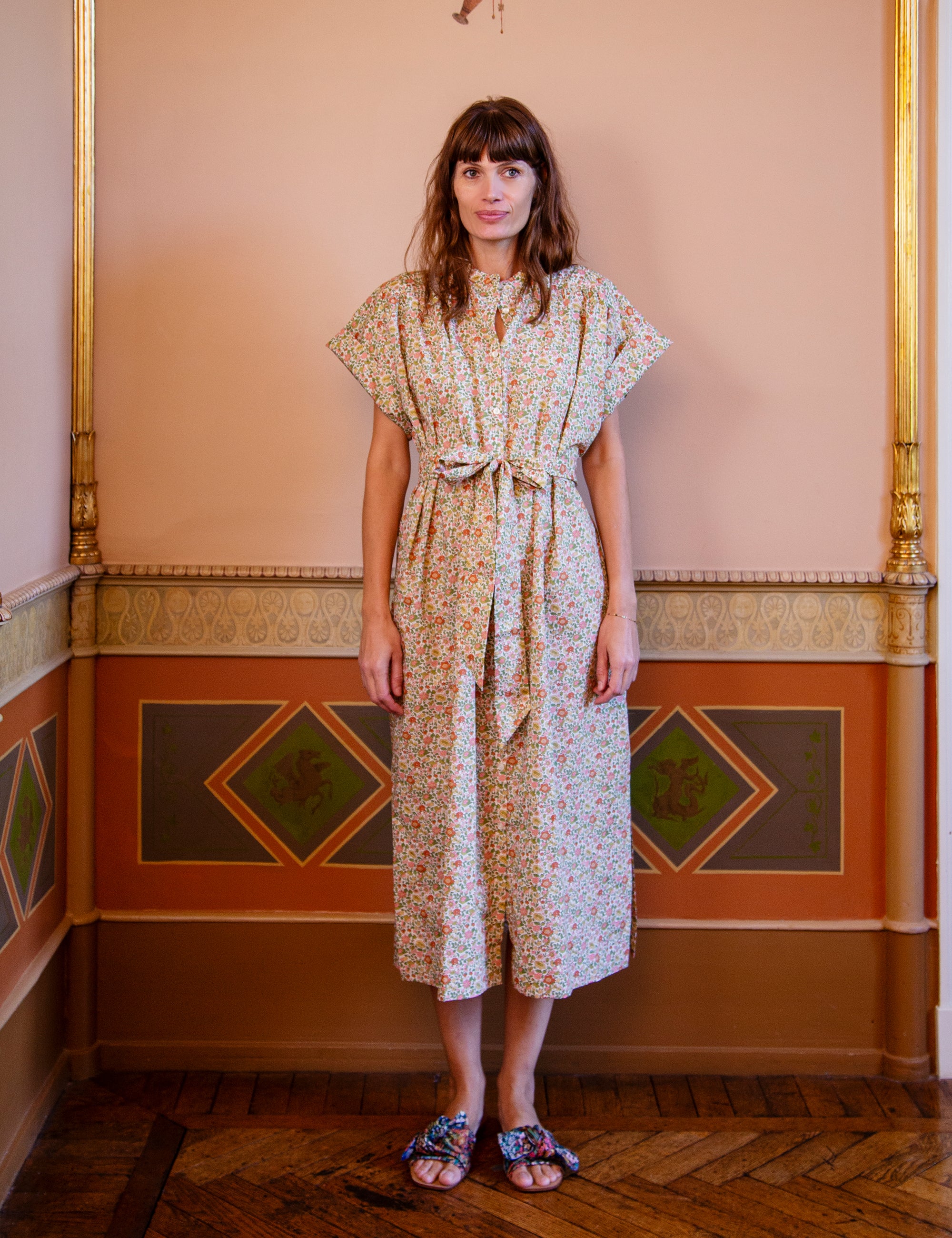 Woman wearing a short sleeved floral dress with belt in an ornate room with decorative walls.
