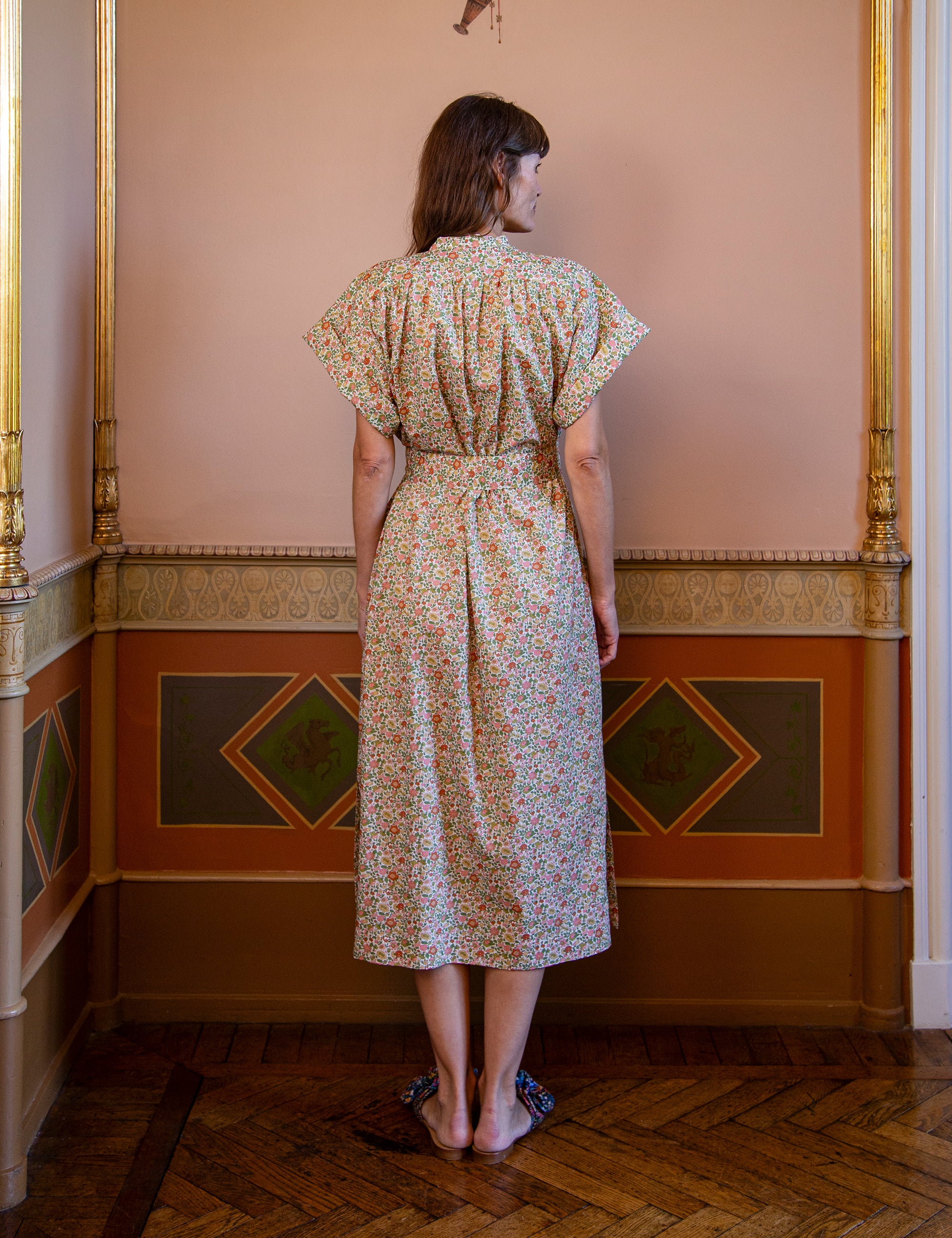 Woman wearing a short sleeved floral dress with belt in an ornate room with decorative walls.