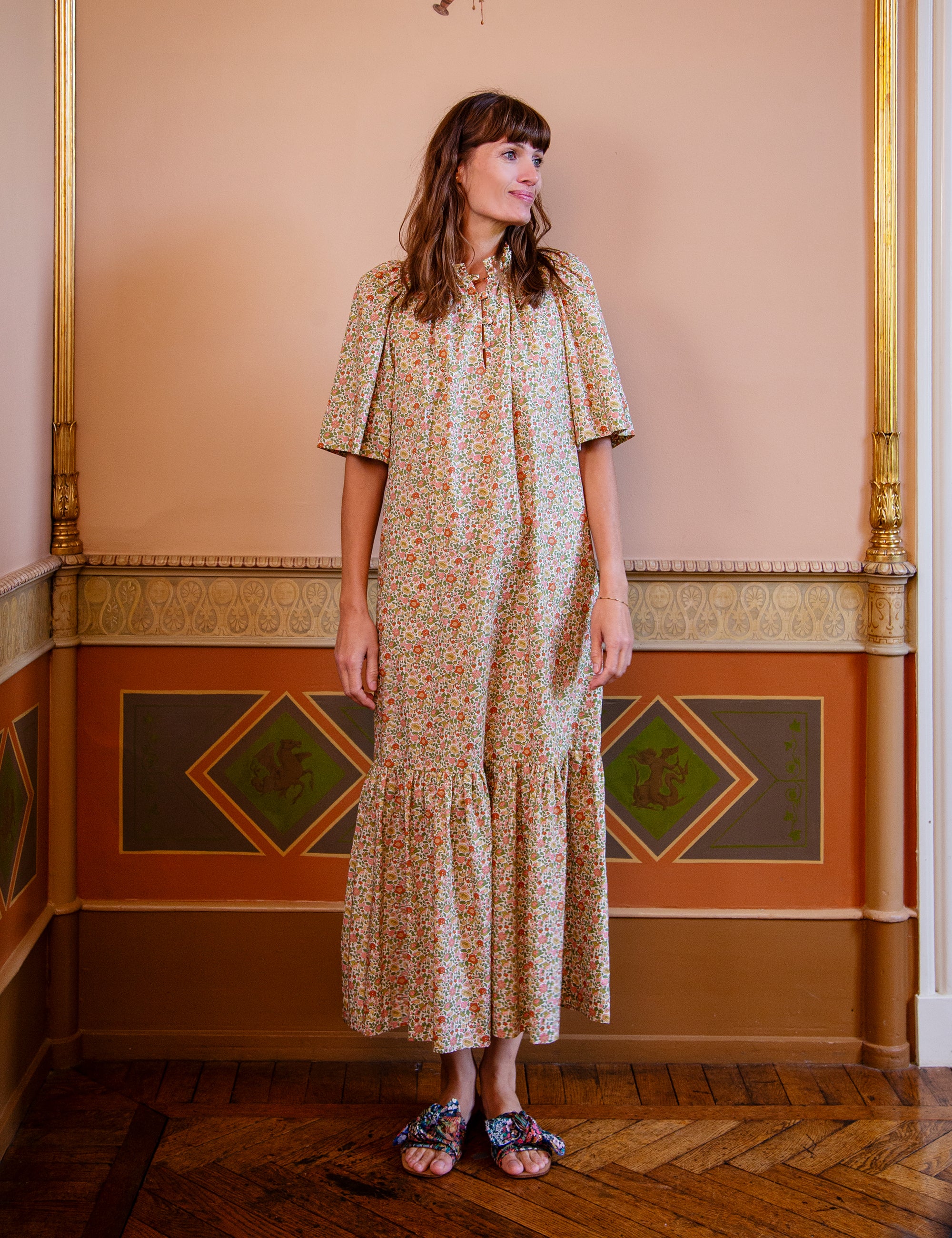 Woman in a floral dress standing in an ornate room with decorative walls.