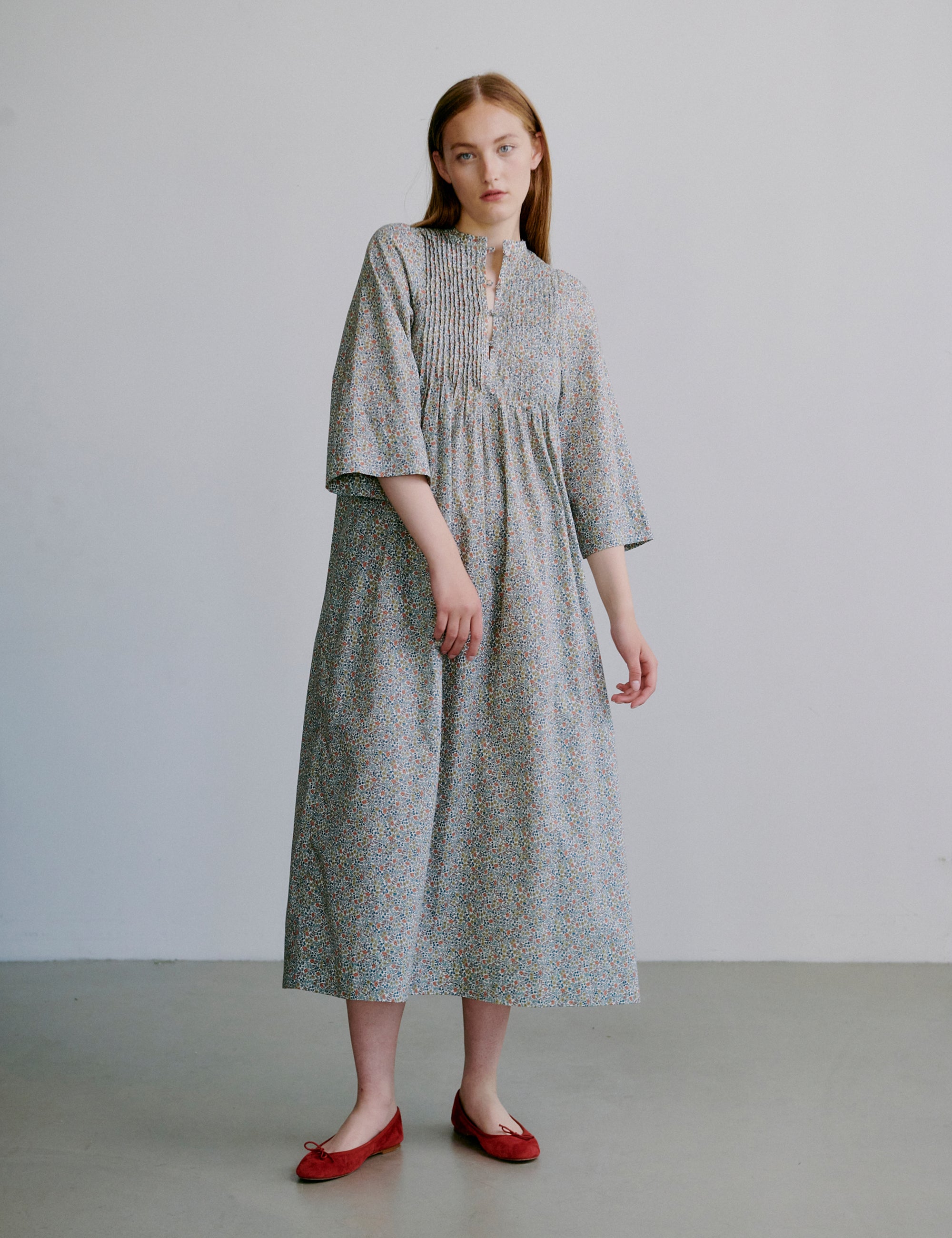 Woman wearing a long, light-colored dress with a subtle floral pattern, semi-long sleeves and red shoes