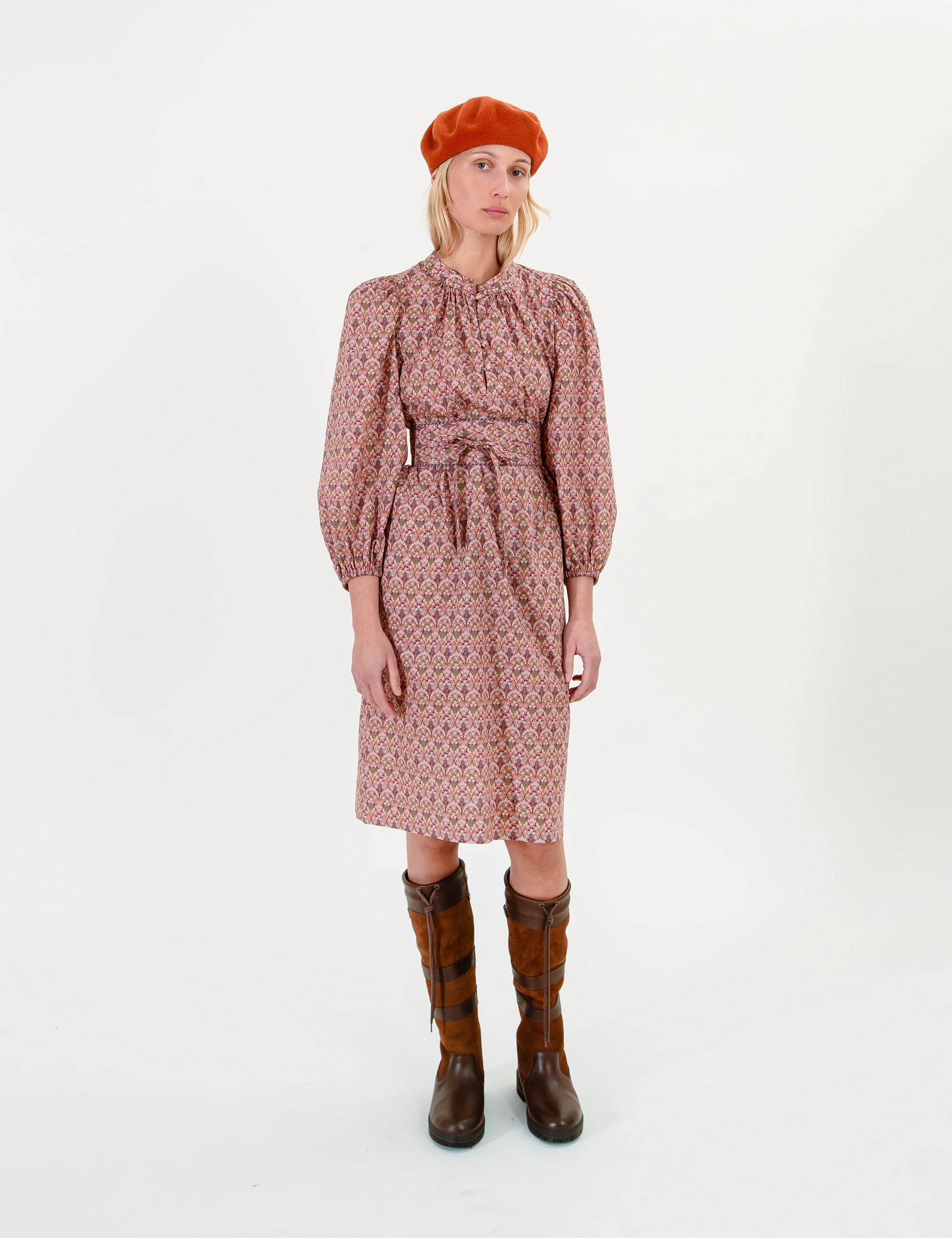 Woman wearing a floral patterned dress with long sleeves and a fabric belt, brown boots, and a red beret on a white background