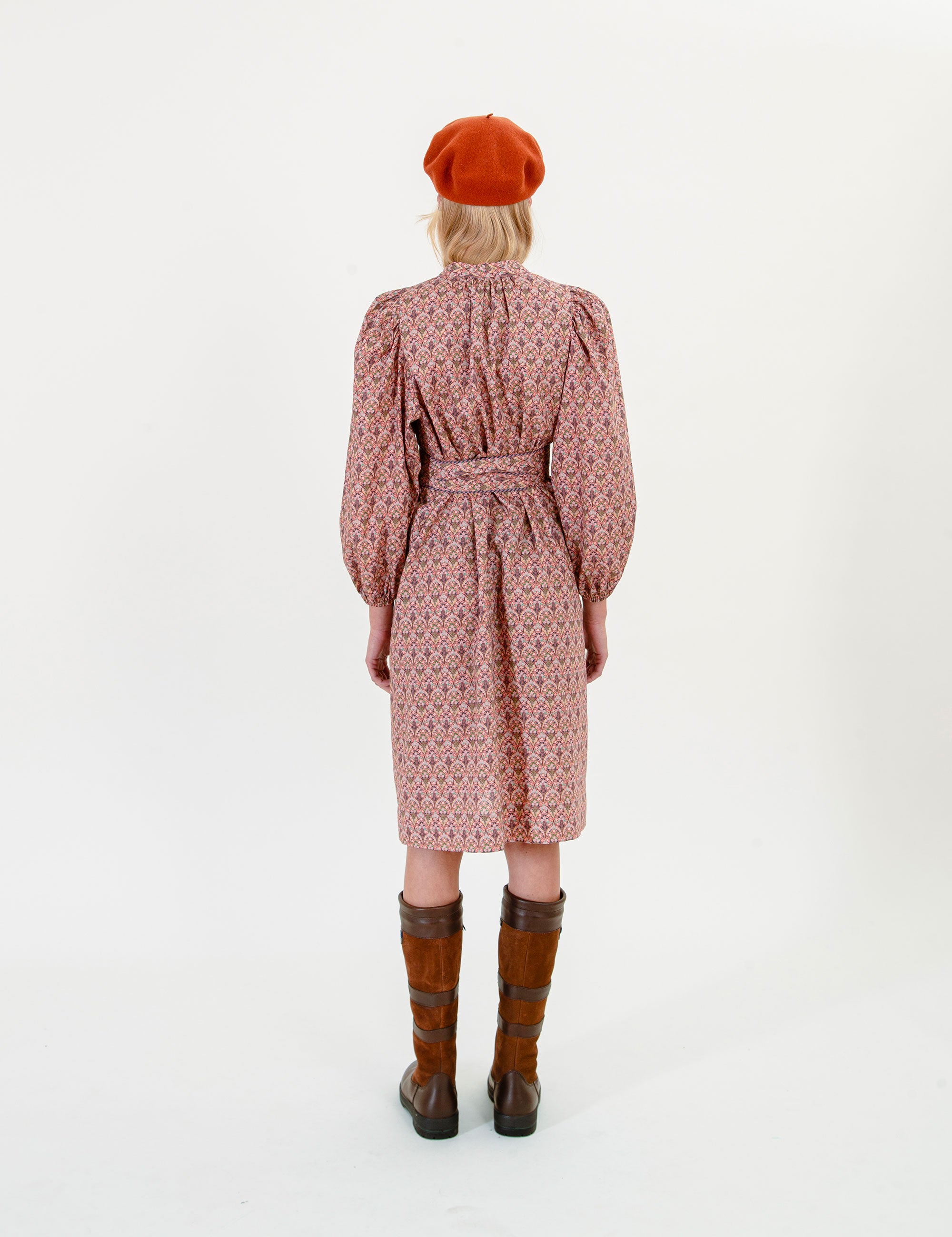 Woman wearing a floral patterned dress with long sleeves and a fabric belt, brown boots, and a red beret on a white background