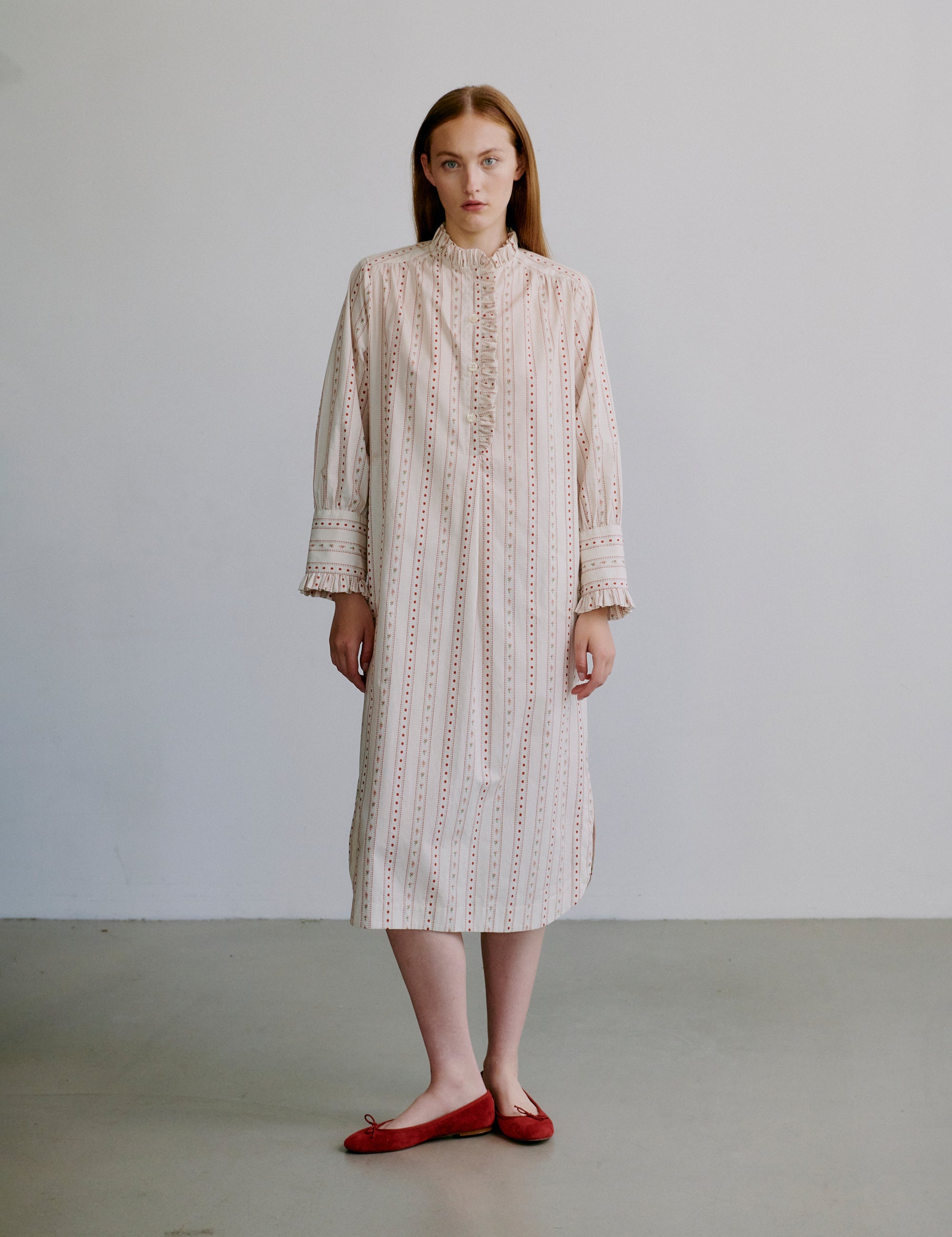 Woman wearing a long, light colored patterned dress with long sleeves and frills against a plain background