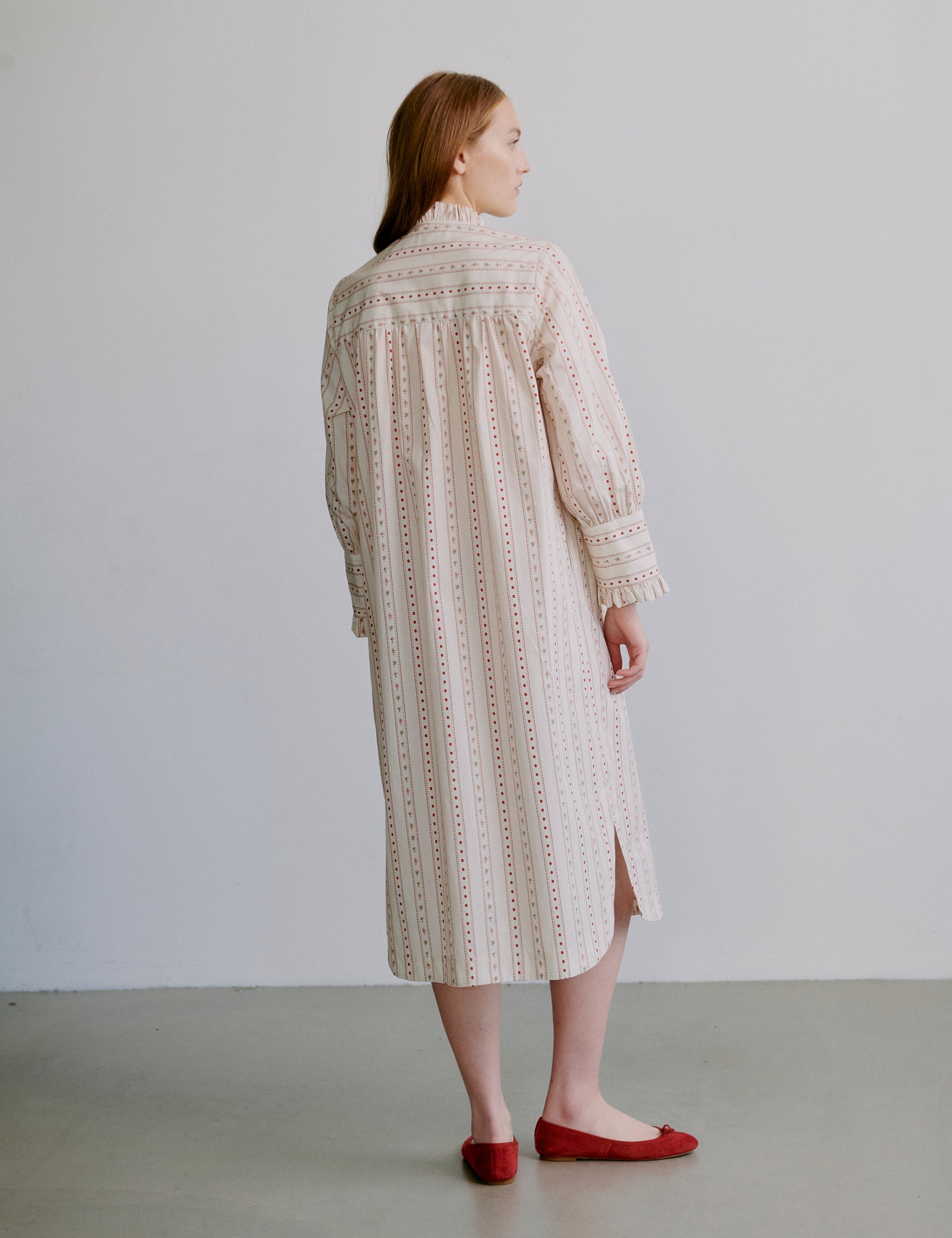 Woman wearing a long, light colored patterned dress with long sleeves and frills against a plain background