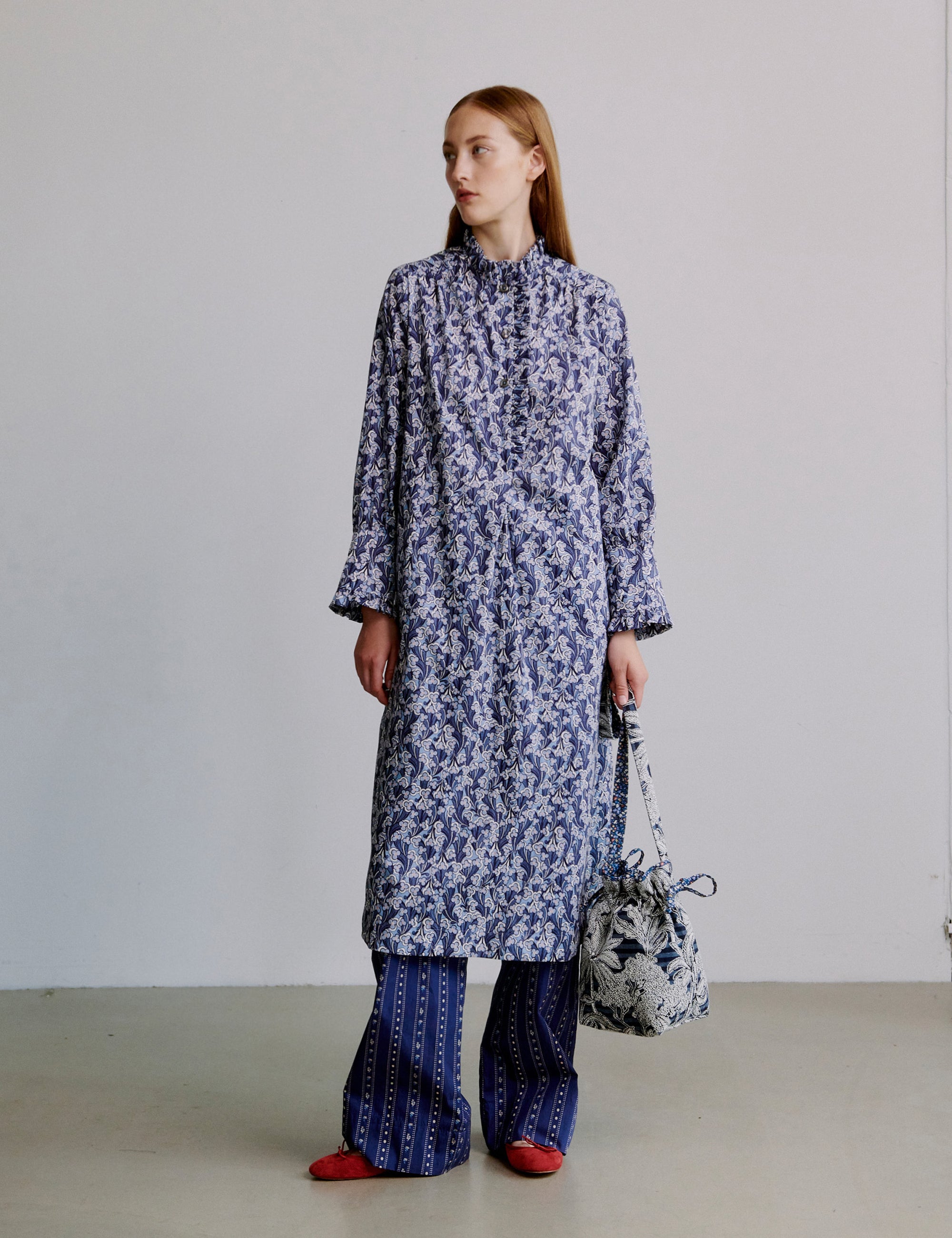 Woman wearing a long blue floral dress with blue striped pants against a plain background holding a floral bag