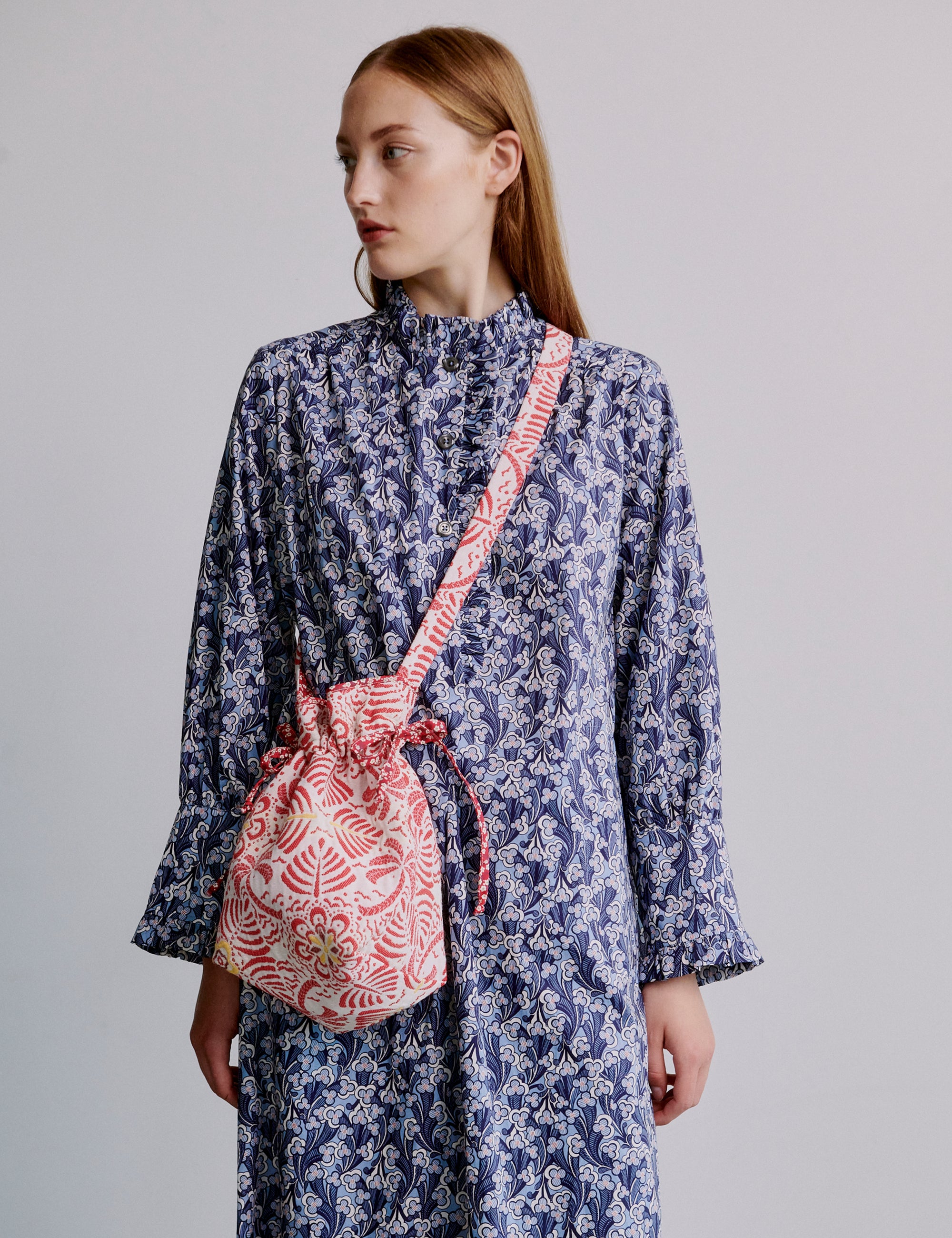 Woman wearing a blue floral dress with a red patterned bag over her shoulder on a plain background