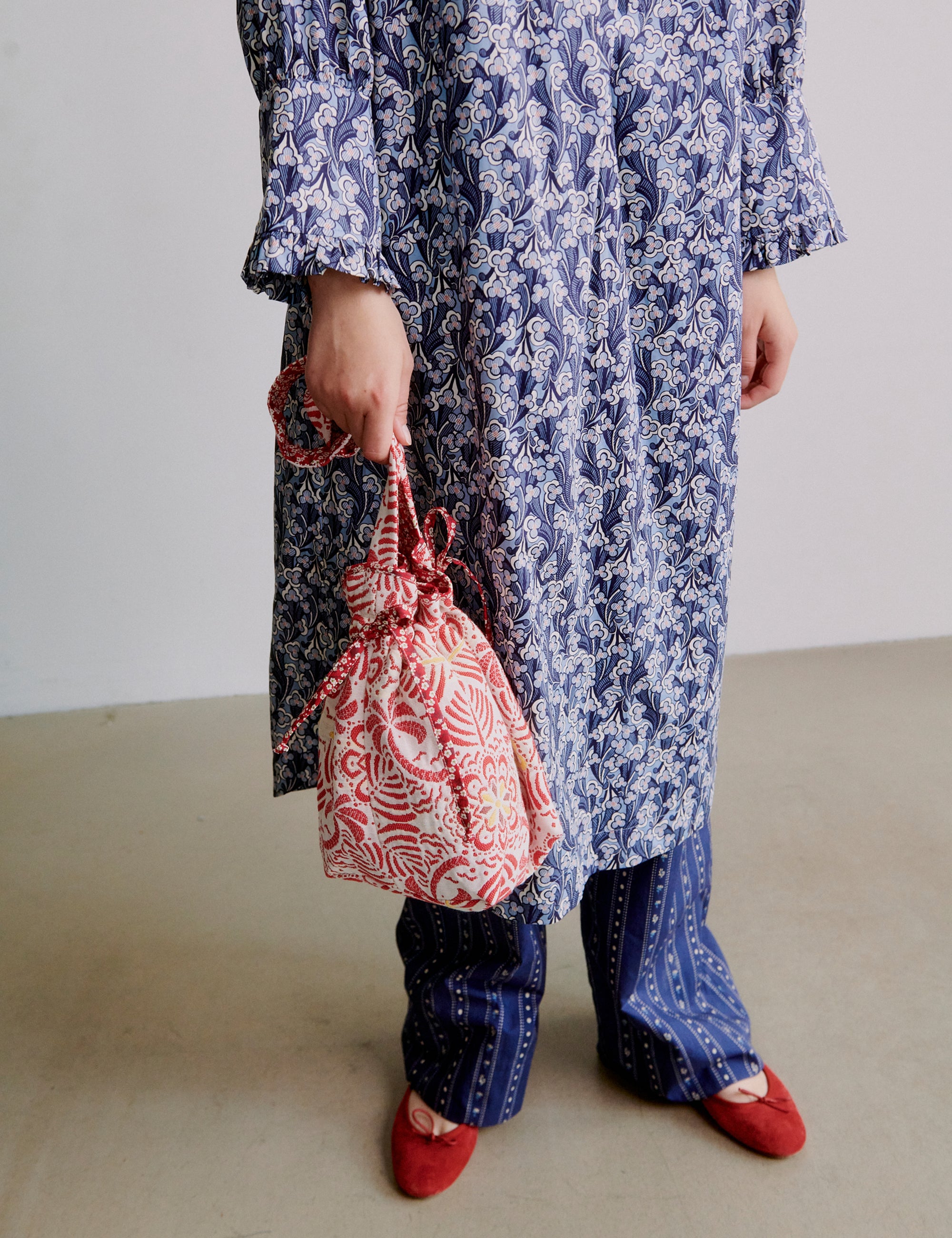 Woman wearing a blue floral dress holding a red patterned bag on a plain background