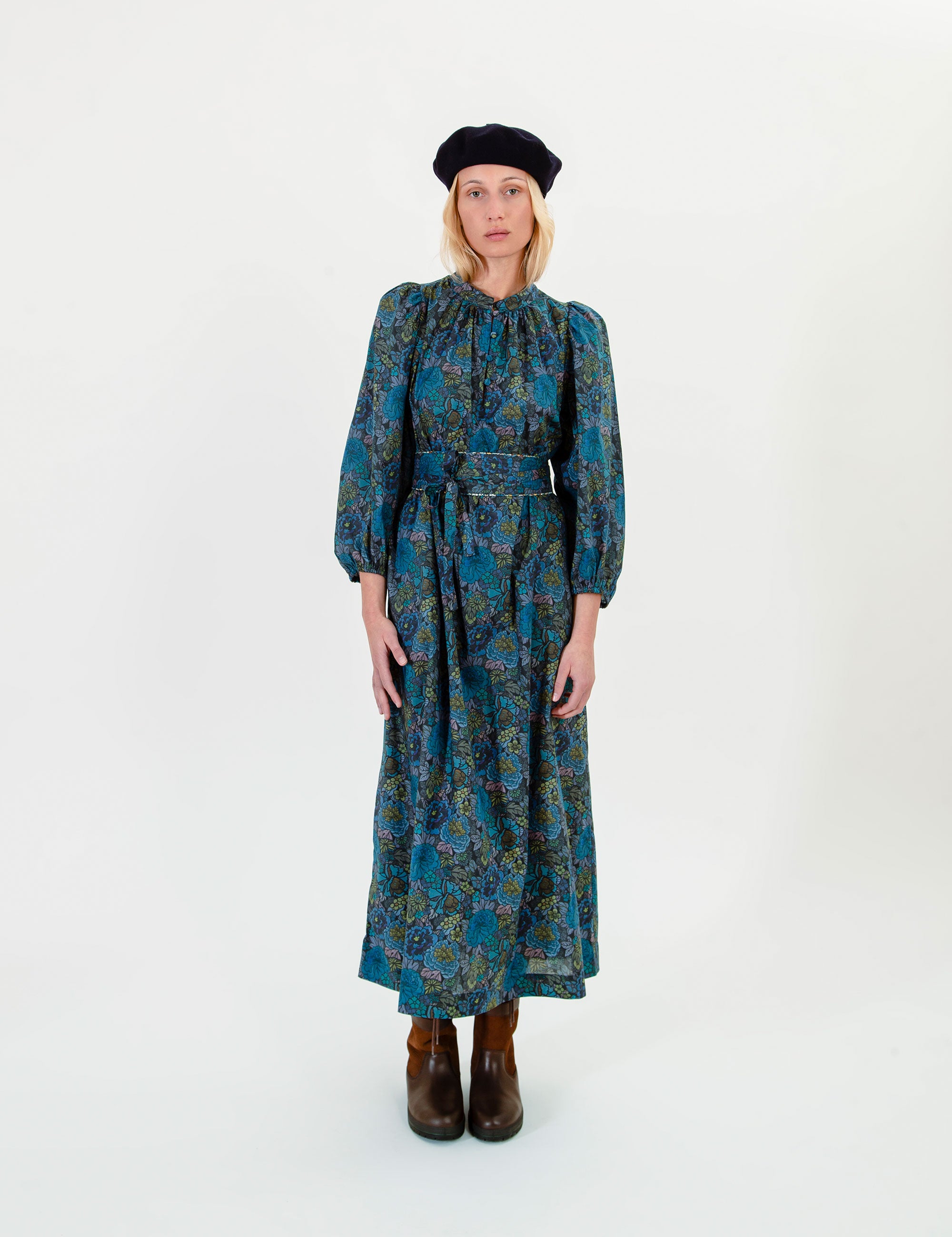 Woman wearing a blue and green floral dress with long sleeves and a belt, standing against a white background.