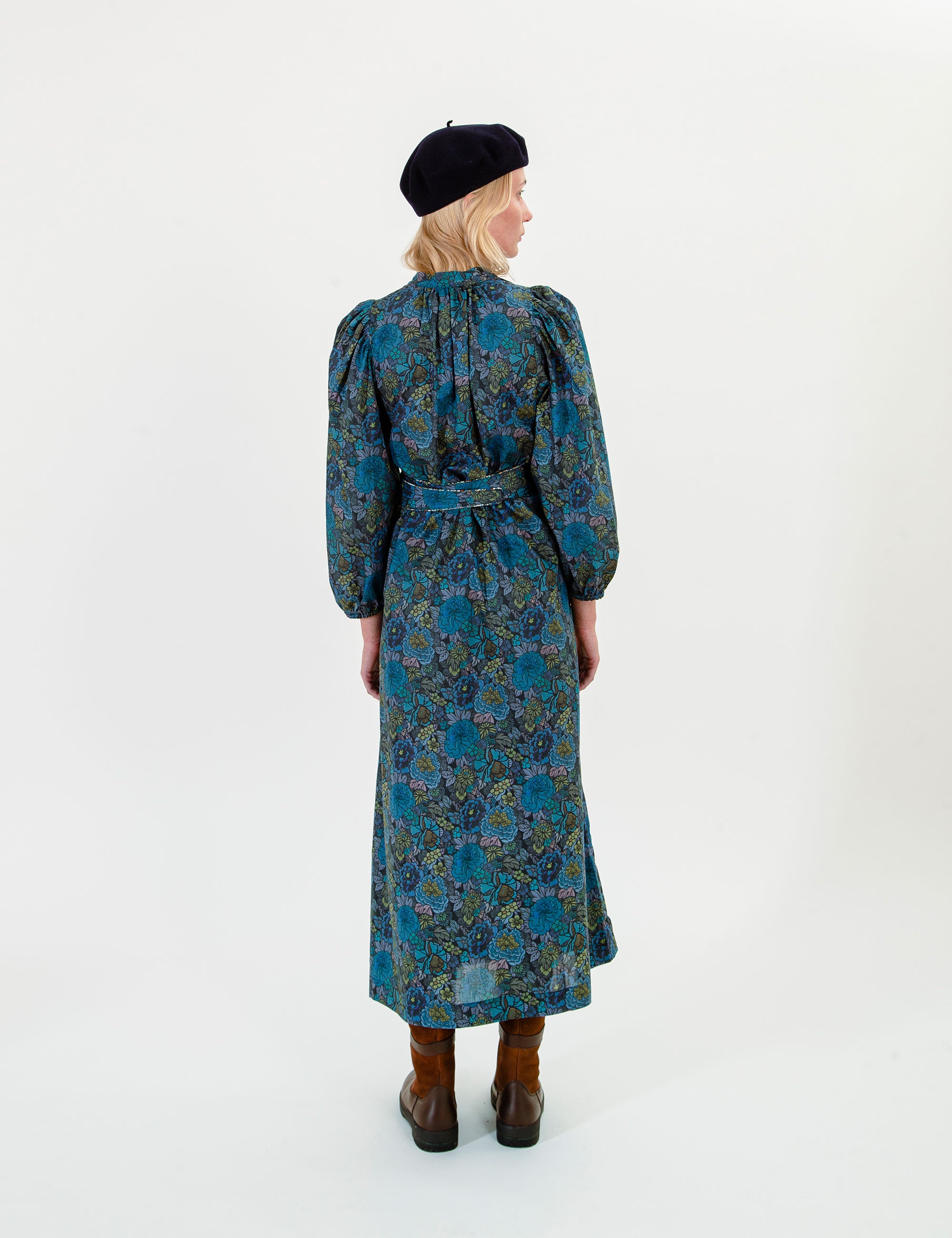 Woman wearing a blue and green floral dress with long sleeves and a belt, standing against a white background.