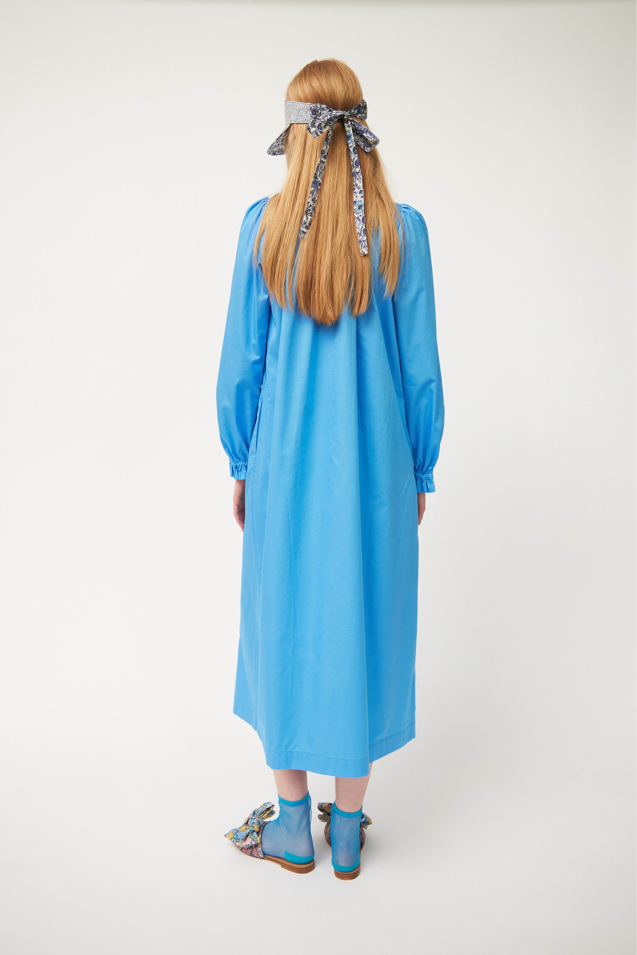 Woman in long blue cotton tunic dress with v-neck, puffed long sleeves and frilled cuffs with sun shade cap and sandals with bows