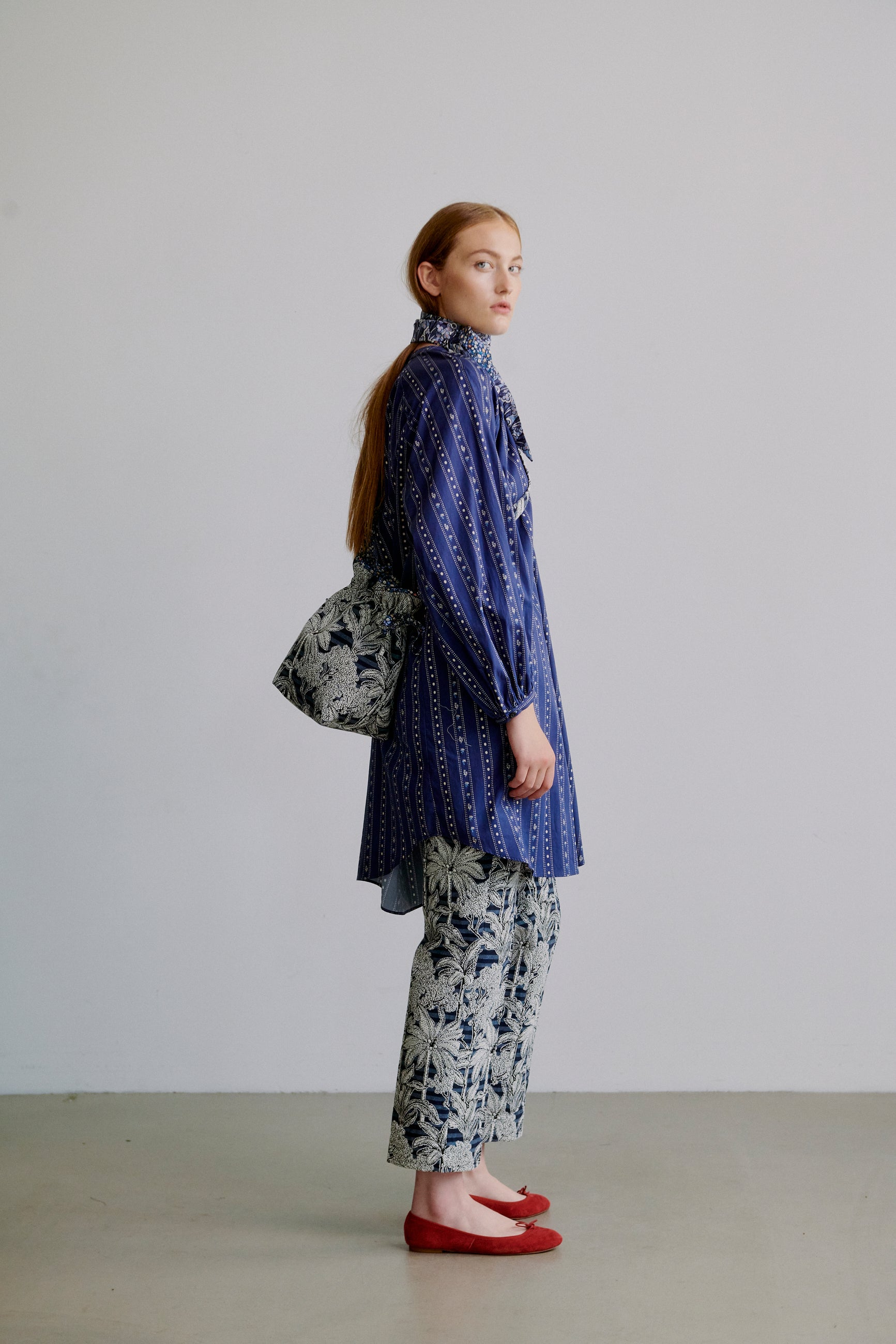 Woman wearing a blue patterned long shirt with long sleeves, blue floral patterned pants, a blue floral scarf around neck and a blue floral fabric bag on the back