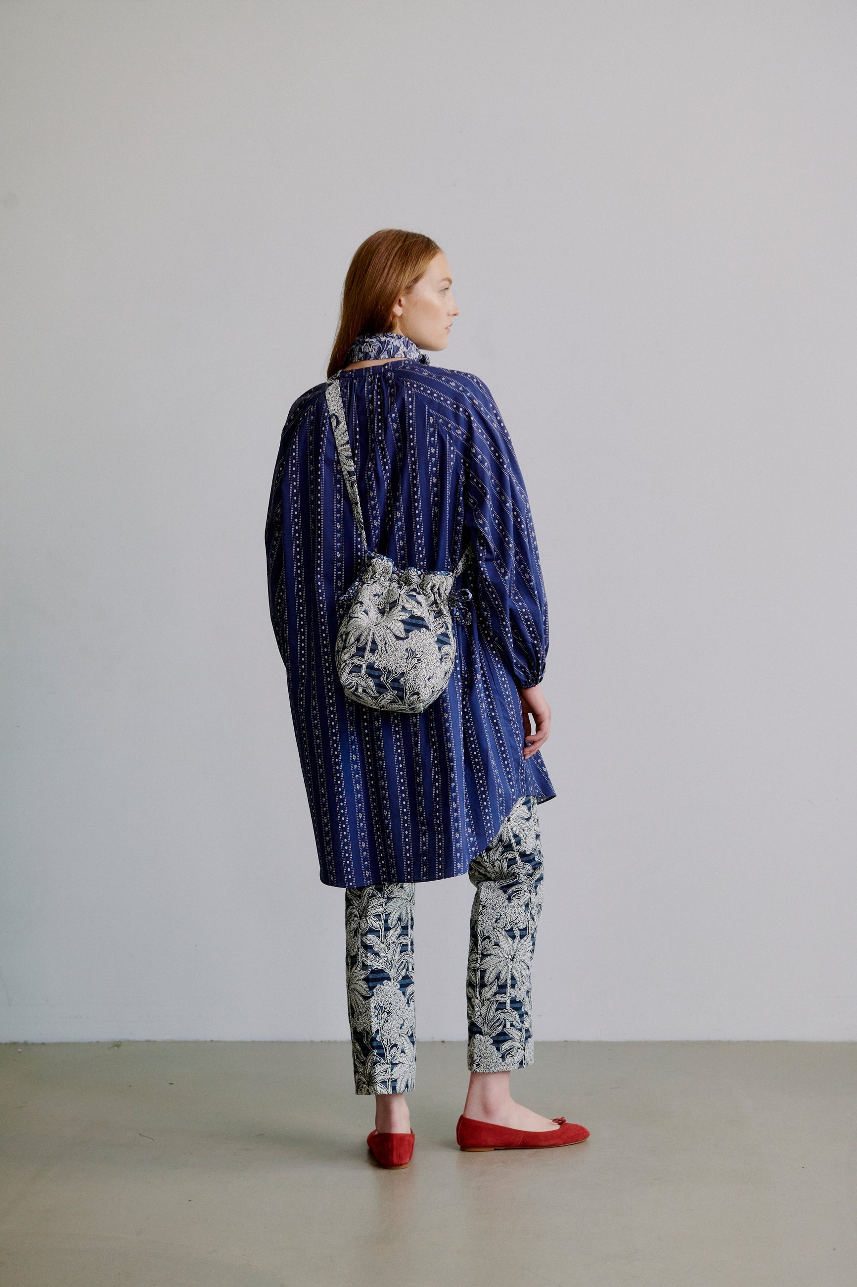 Woman wearing a blue patterned long shirt with long sleeves, blue floral patterned pants, a blue floral scarf around neck and a blue floral fabric bag on the back