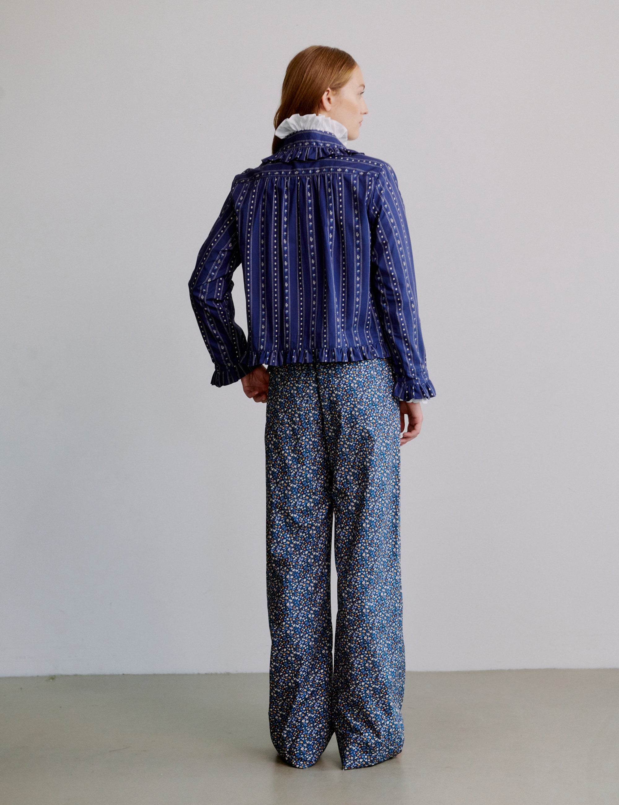 Woman wearing a blue patterned shirt with frill collar and floral patterned blue pants on a plain background