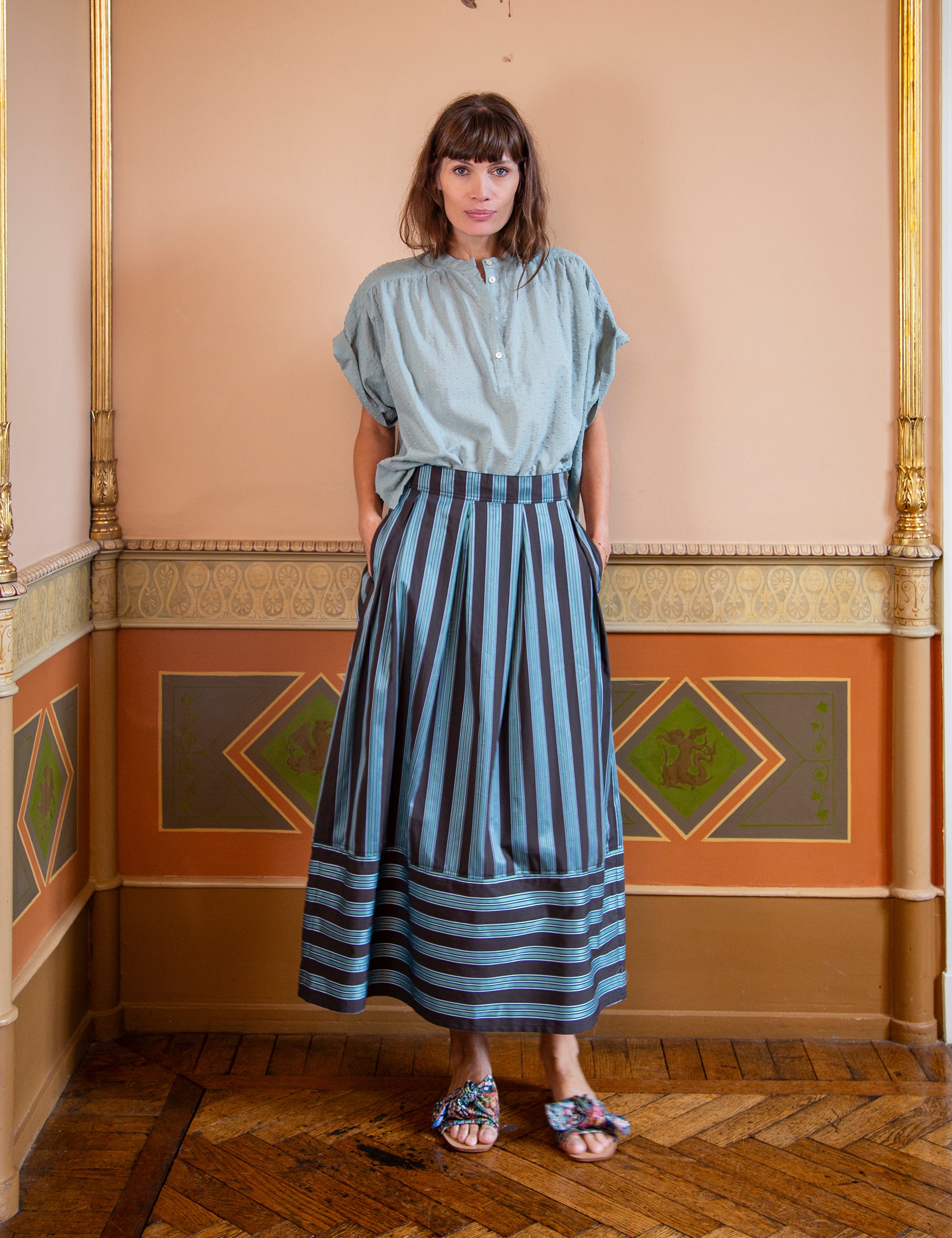 Woman wearing a light green top and striped maxi skirt standing in an ornate room.