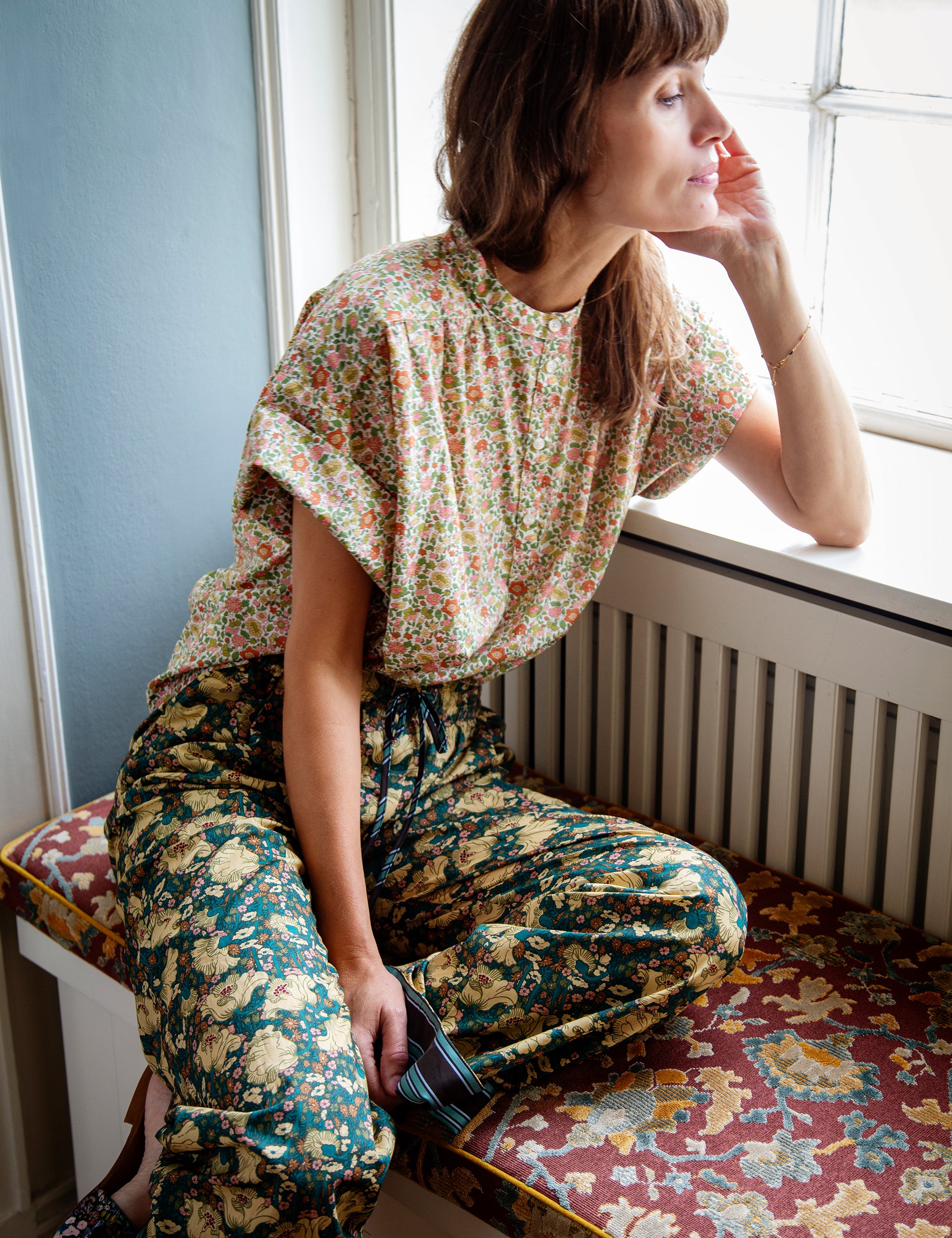 Woman sitting by a window wearing a floral top and patterned pants.